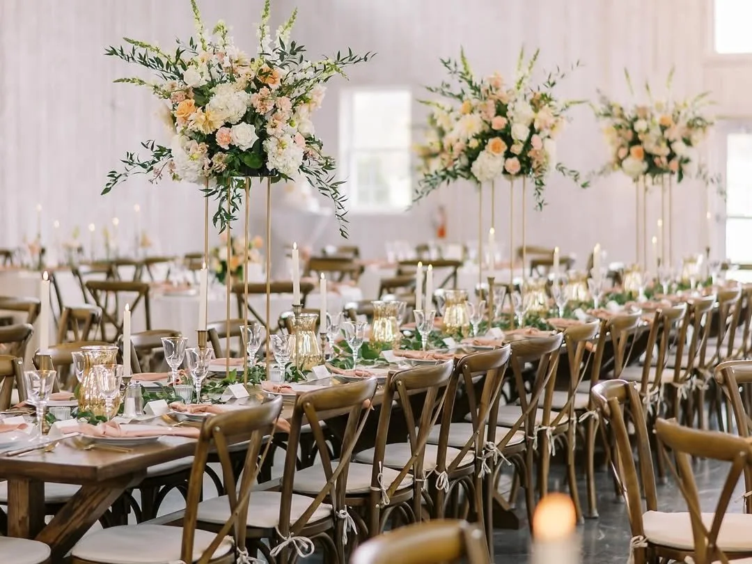 Elegant event reception with long wooden tables decorated with large floral centerpieces, candleholders, glassware, and pastel-colored napkins in a bright, airy venue.