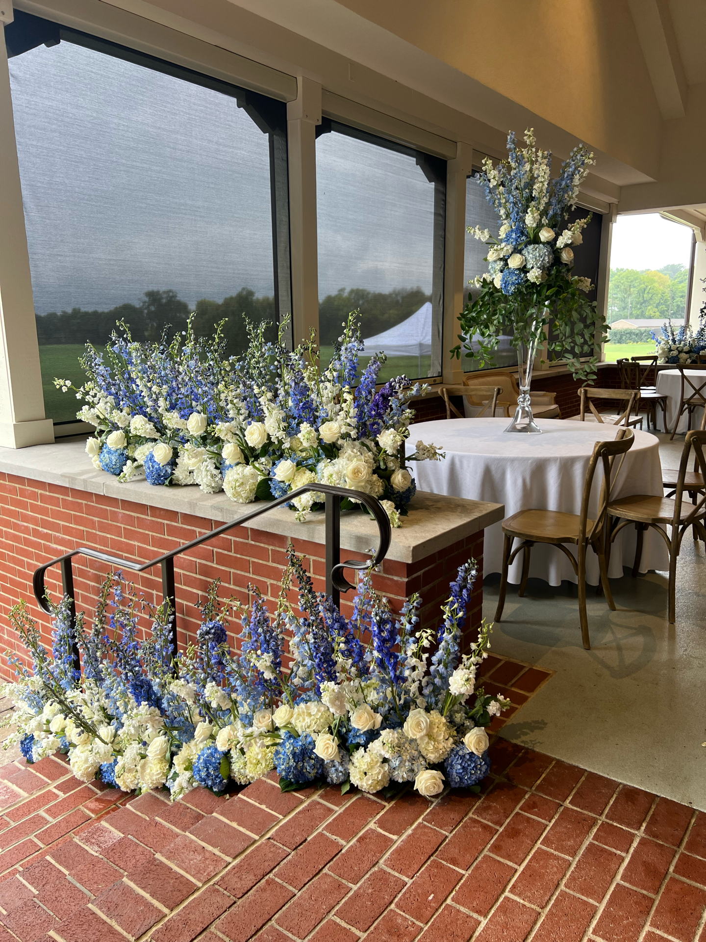 Indoor event space decorated with floral arrangements of white and blue flowers on tables and a brick ledge, with large windows showing a green outdoor landscape.