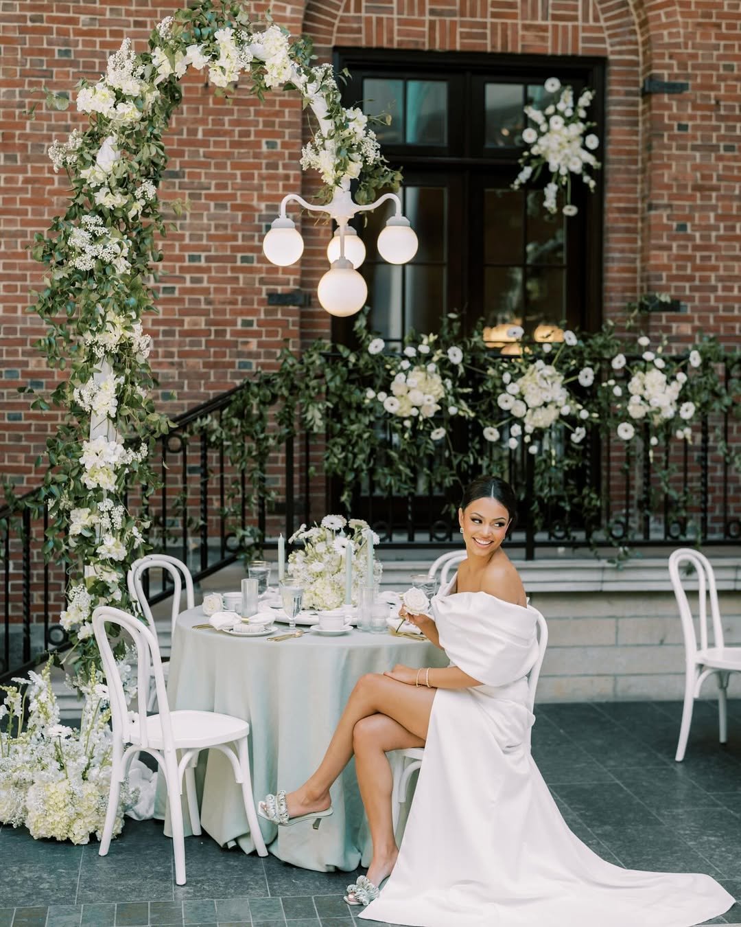 A woman in a white dress sitting at an elegant outdoor table decorated with white flowers and greenery. The setting features a brick building with a decorated arch, black iron railing, and large windows, with soft lighting from a chandelier overhead.