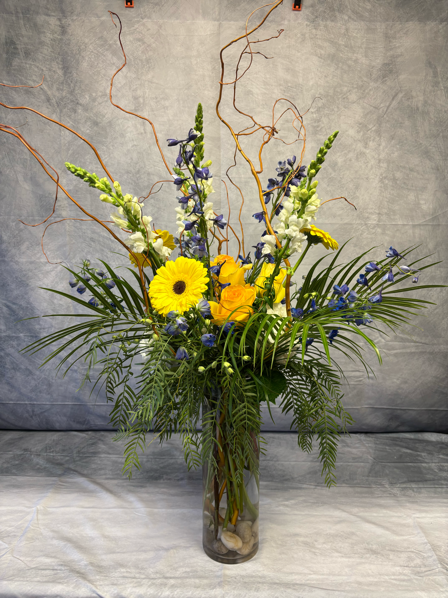 A vibrant flower arrangement in a glass vase featuring yellow, purple, and white flowers, surrounded by greenery and decorative branches on a neutral background.