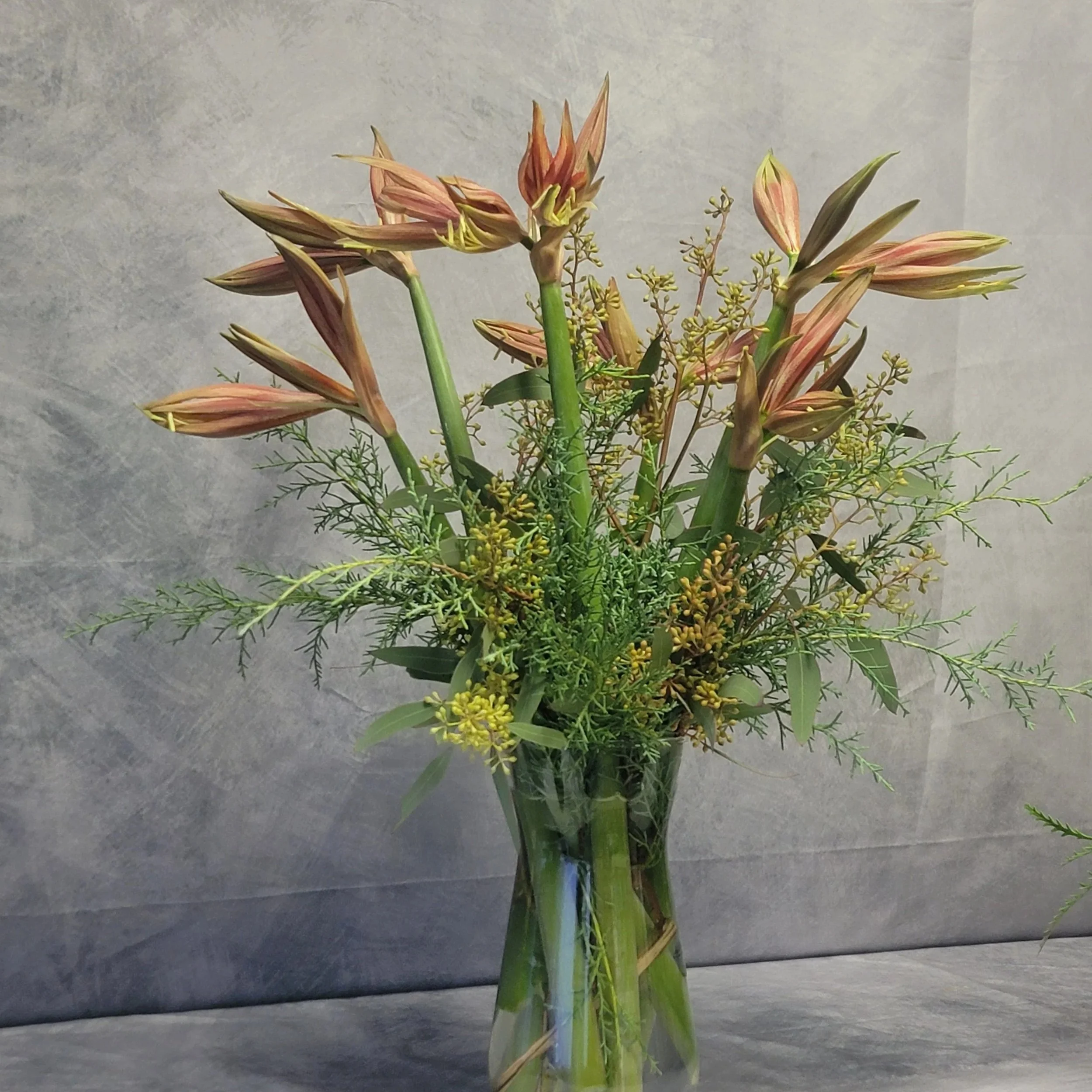 A tall clear glass vase holding a bouquet of pinkish-orange lilies and green foliage, placed on a gray surface with a gray background.