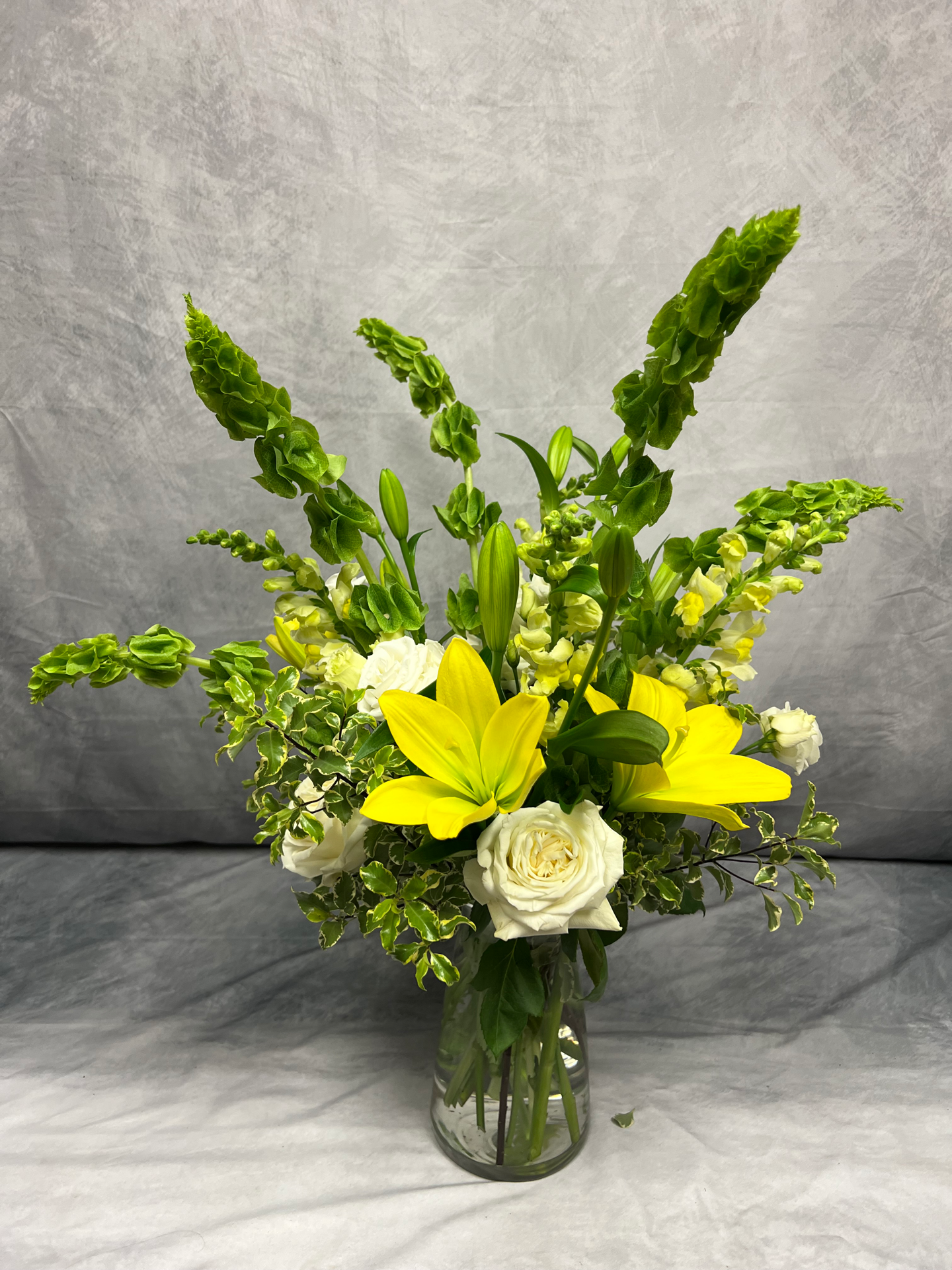 A floral arrangement in a glass vase featuring white roses, yellow lilies, white snapdragons, and assorted green foliage, set against a gray background.