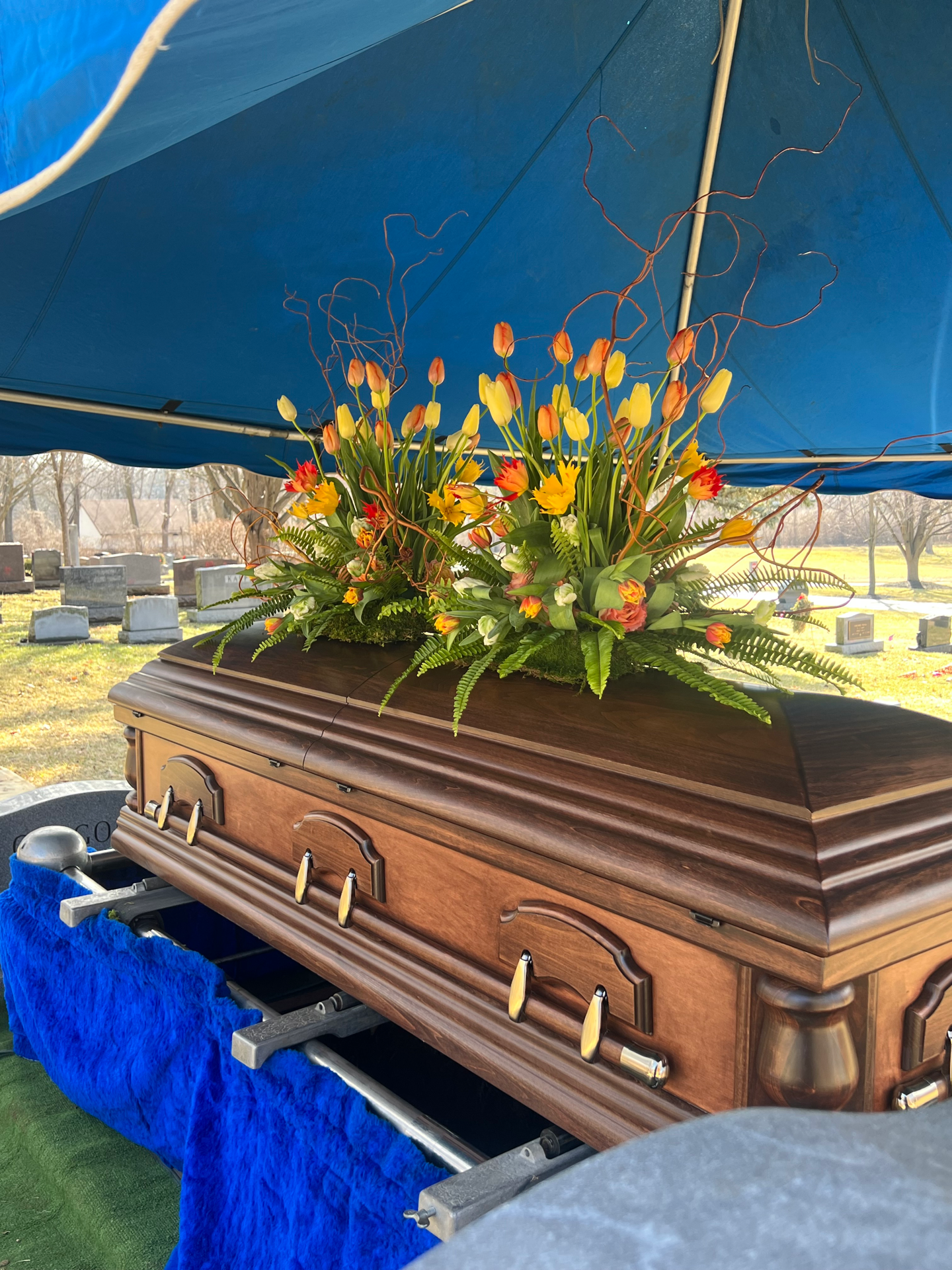 A wooden casket with a floral arrangement of yellow, orange, and white flowers and green foliage on top, set inside a blue tent at a cemetery.