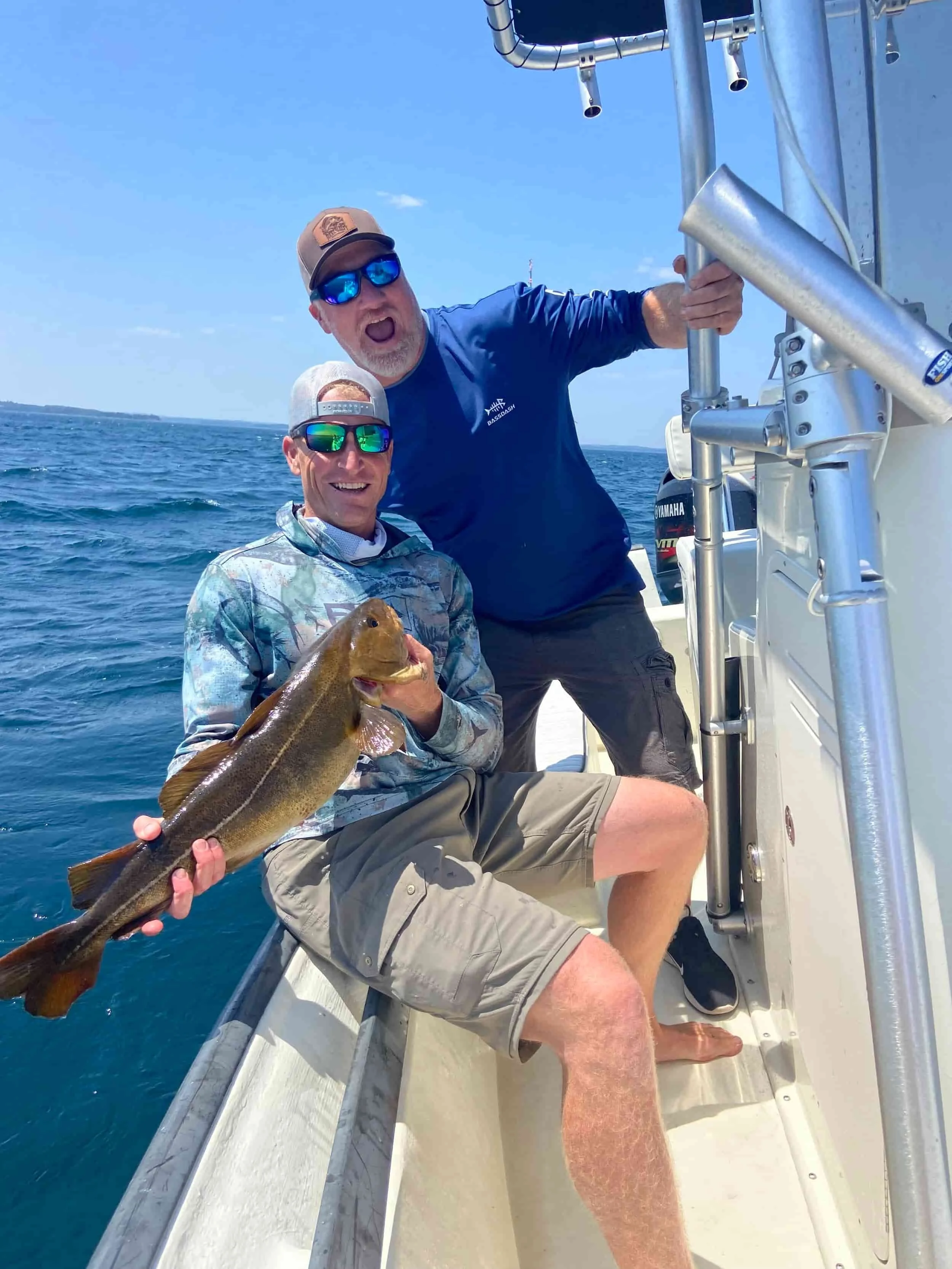 Two men on a boat, one holding a fish, celebrating a fishing catch, with blue sky and ocean in the background.