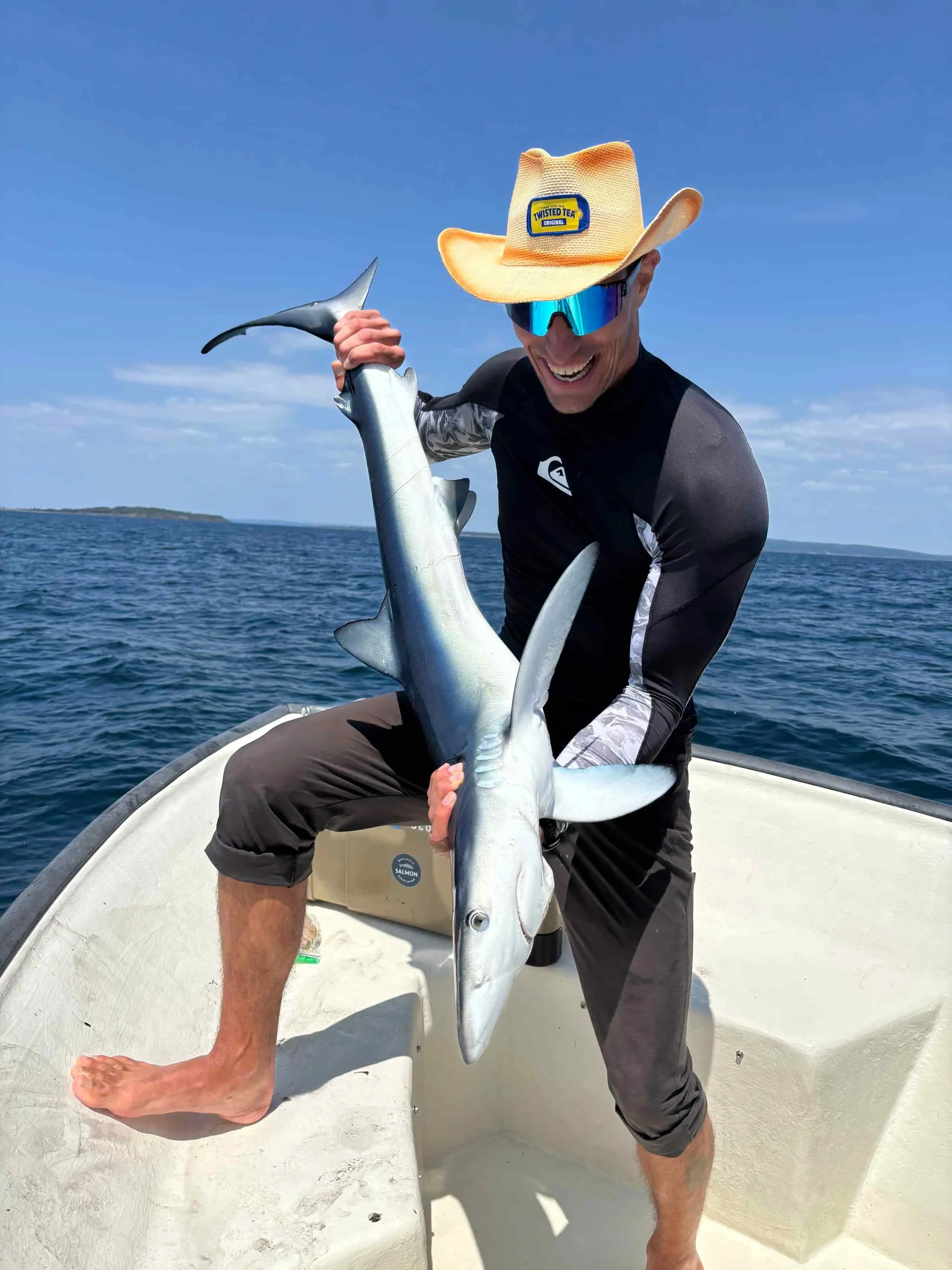 Smiling man wearing a straw cowboy hat and sunglasses holding a large shark on a boat in the ocean.
