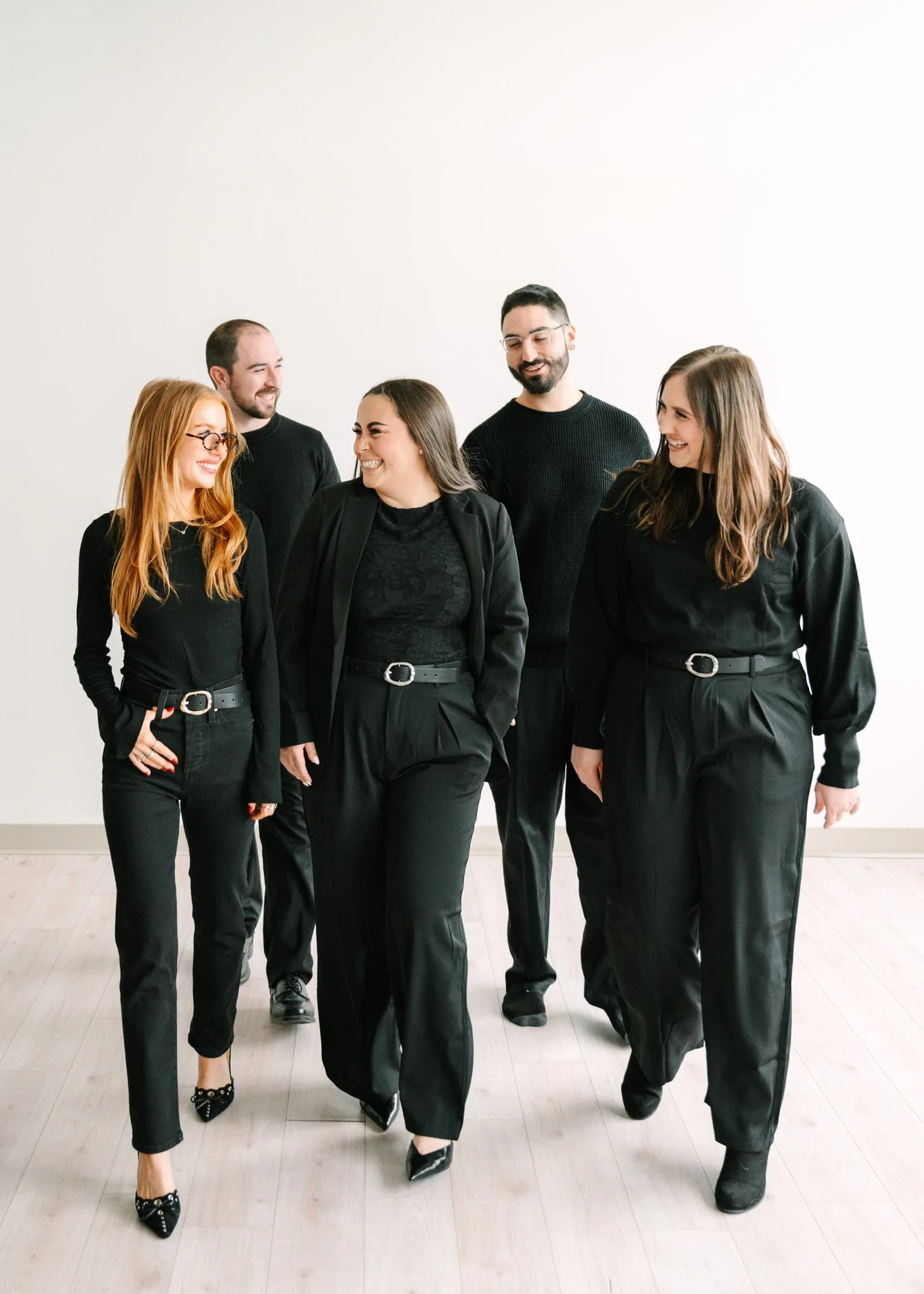 Group of five diverse young adults walking together in casual black outfits against a plain white background, smiling and engaging with each other.