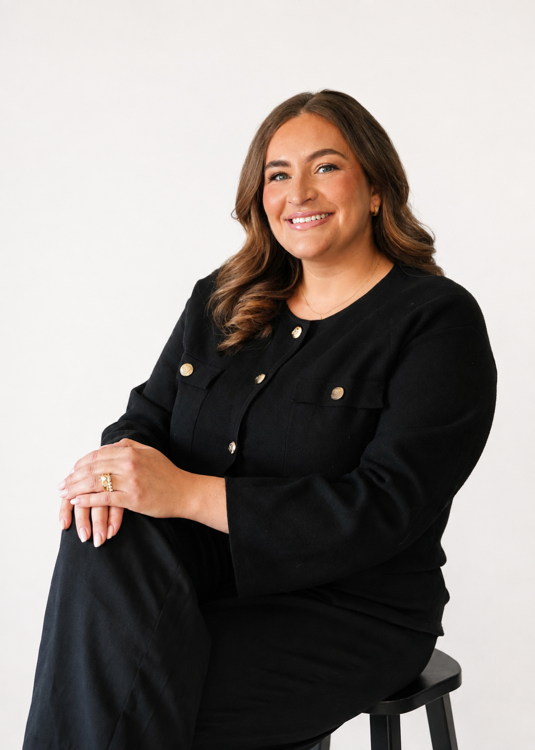 A woman sitting on a black stool, wearing a black jacket with gold buttons, smiling, with long wavy brown hair and light makeup against a plain white background.