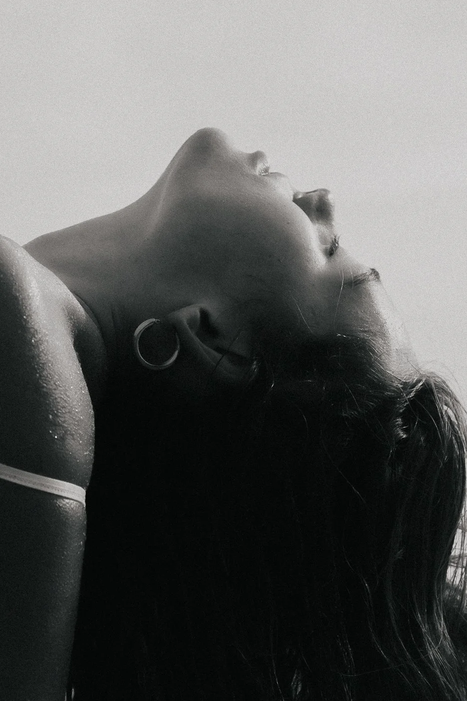 Black and white photo of a woman with wet hair, her head tilted back, showing her profile, wearing a hoop earring and a shoulder strap.