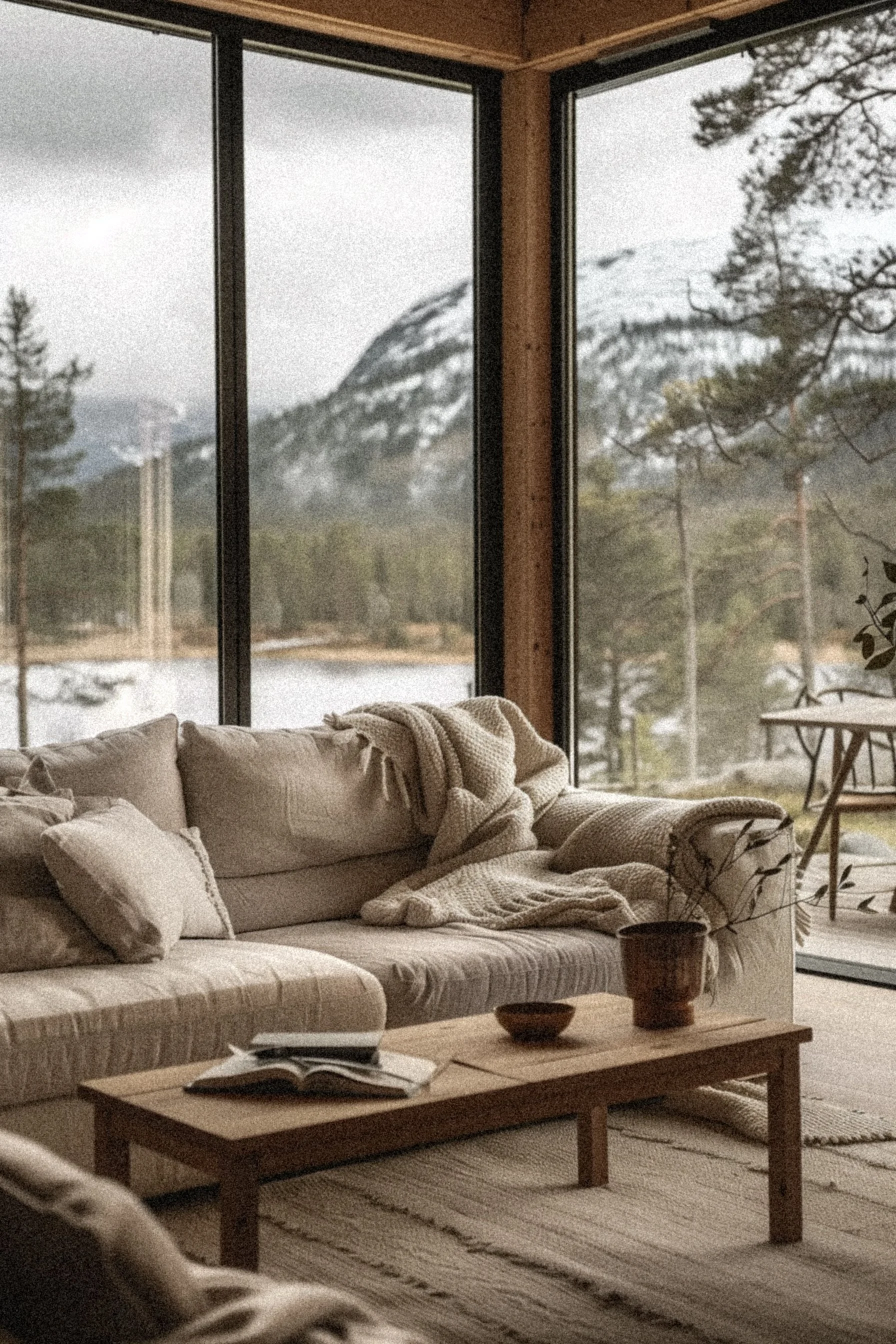 Living room with a beige sofa, throw blanket, and pillows, next to large glass windows showing a mountain and forest landscape outside.