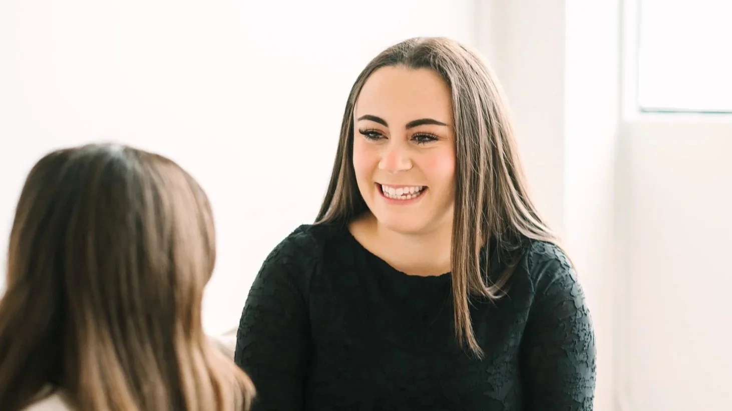 Two women engaging in a friendly conversation, one with long dark hair smiling warmly, wearing a black lace top, indoors with bright natural lighting.