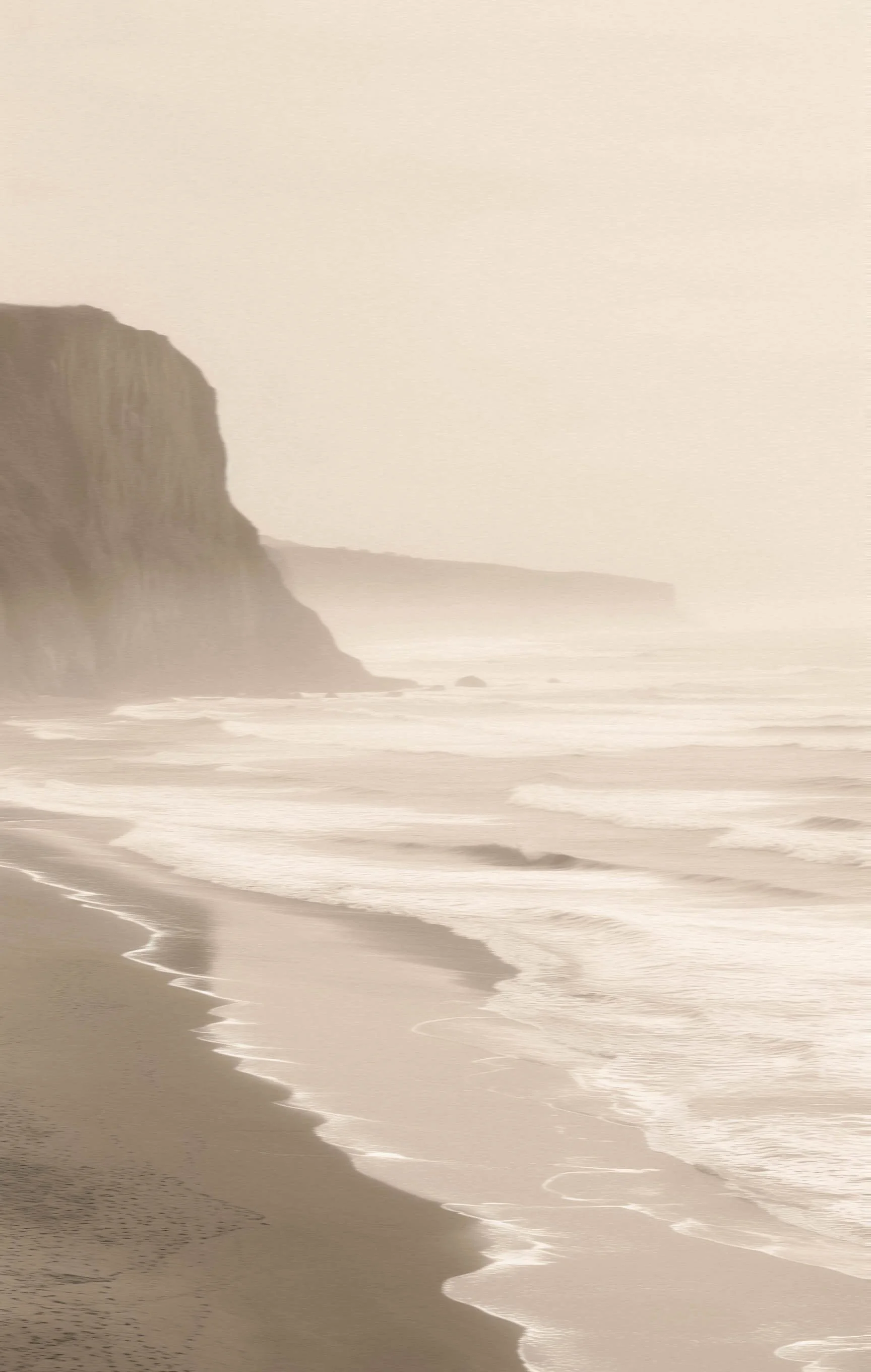A foggy beach scene with cliffs on the left and waves gently rolling onto the shore.