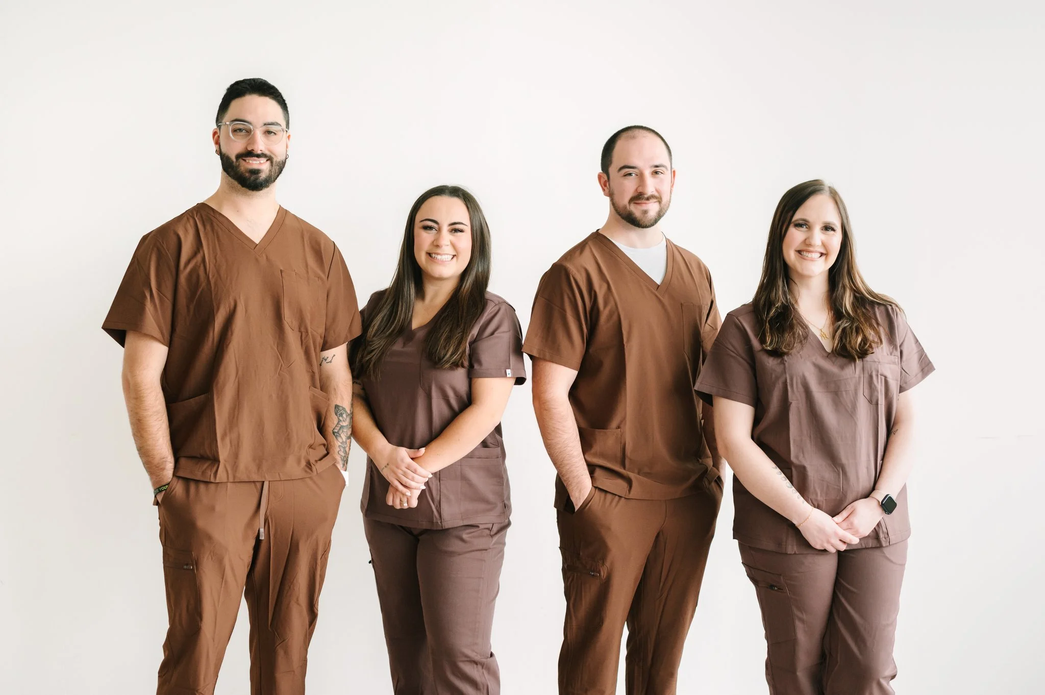 A group of four healthcare professionals standing against a plain white background, wearing brown scrubs, smiling for the camera.