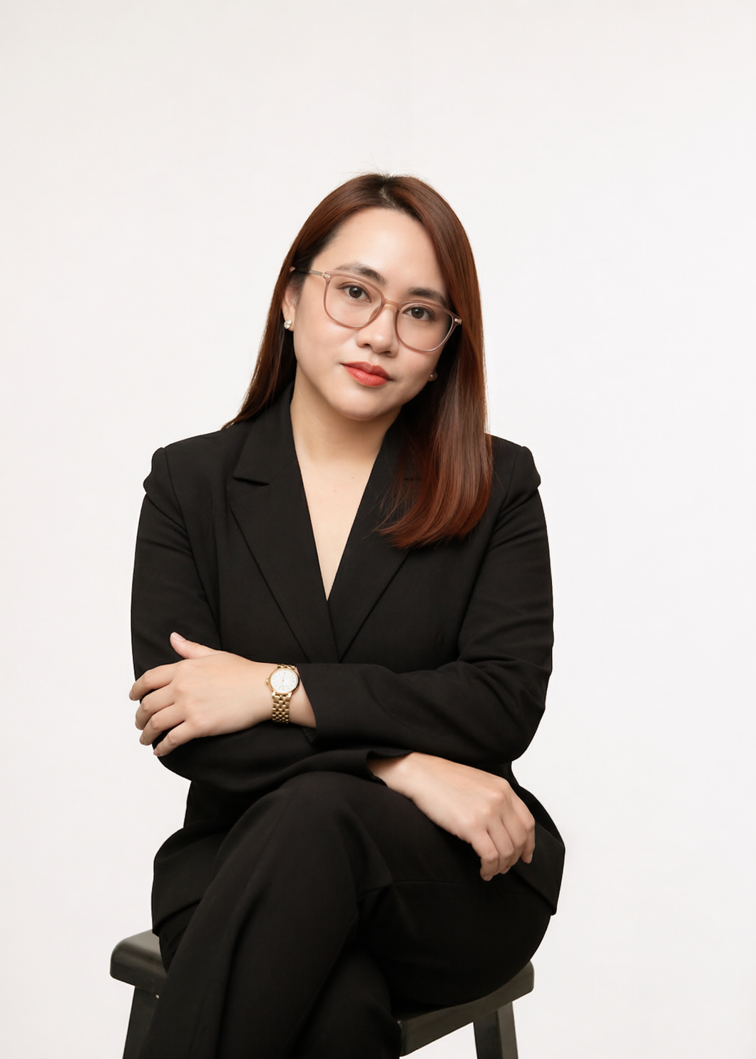 A woman with shoulder-length brown hair, glasses, and a black blazer sitting cross-legged on a stool against a plain white background.