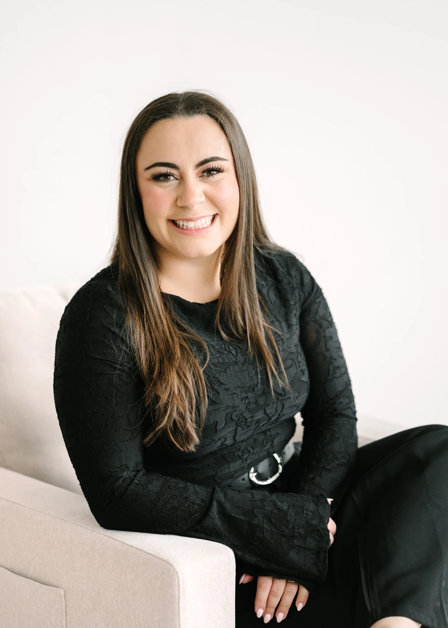 A woman with long brown hair and a black lace long-sleeve top, smiling and sitting on a light-colored sofa against a plain white background.