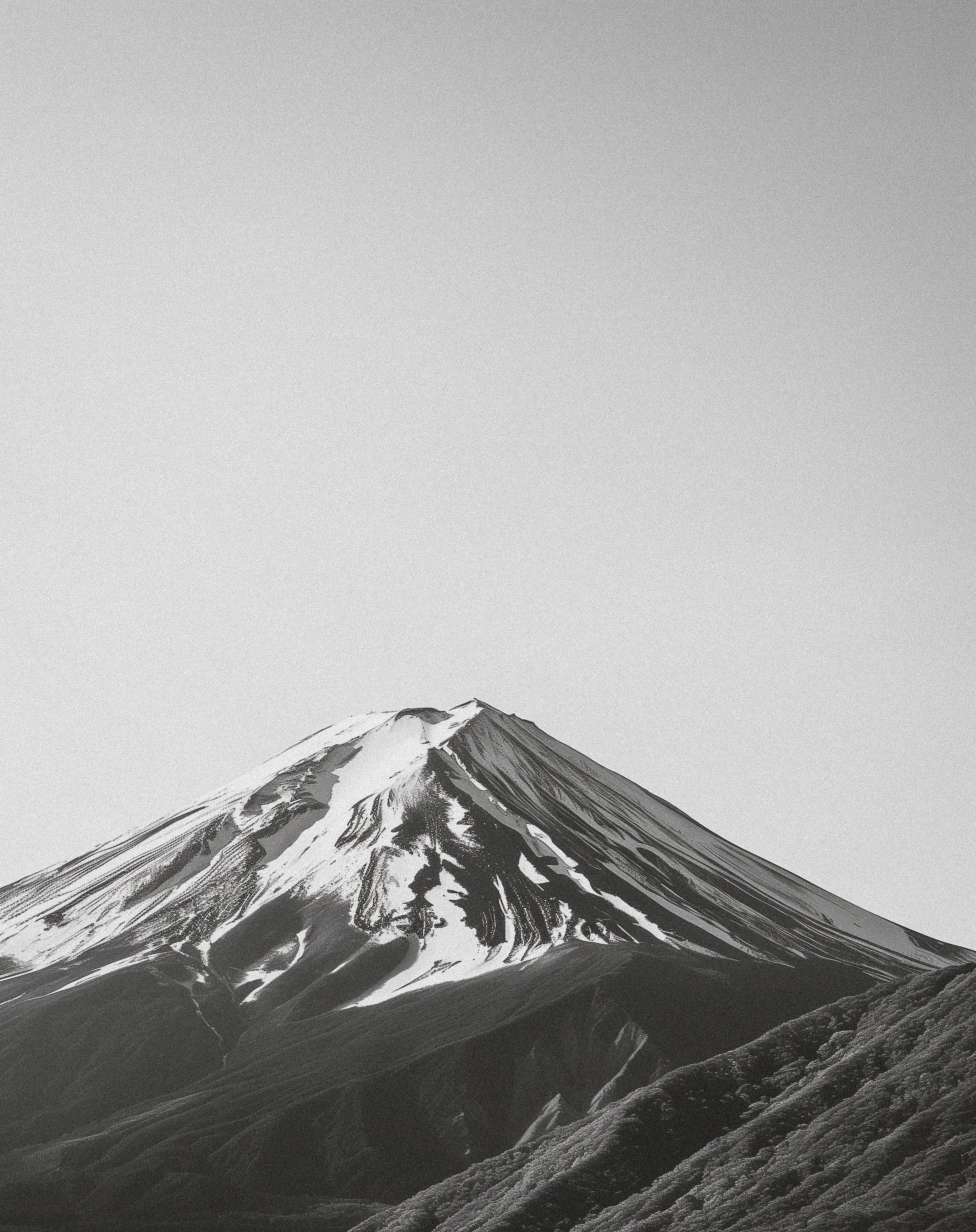 Black and white photograph of a snow-capped mountain peak, likely Mount Fuji, with surrounding rugged terrain.
