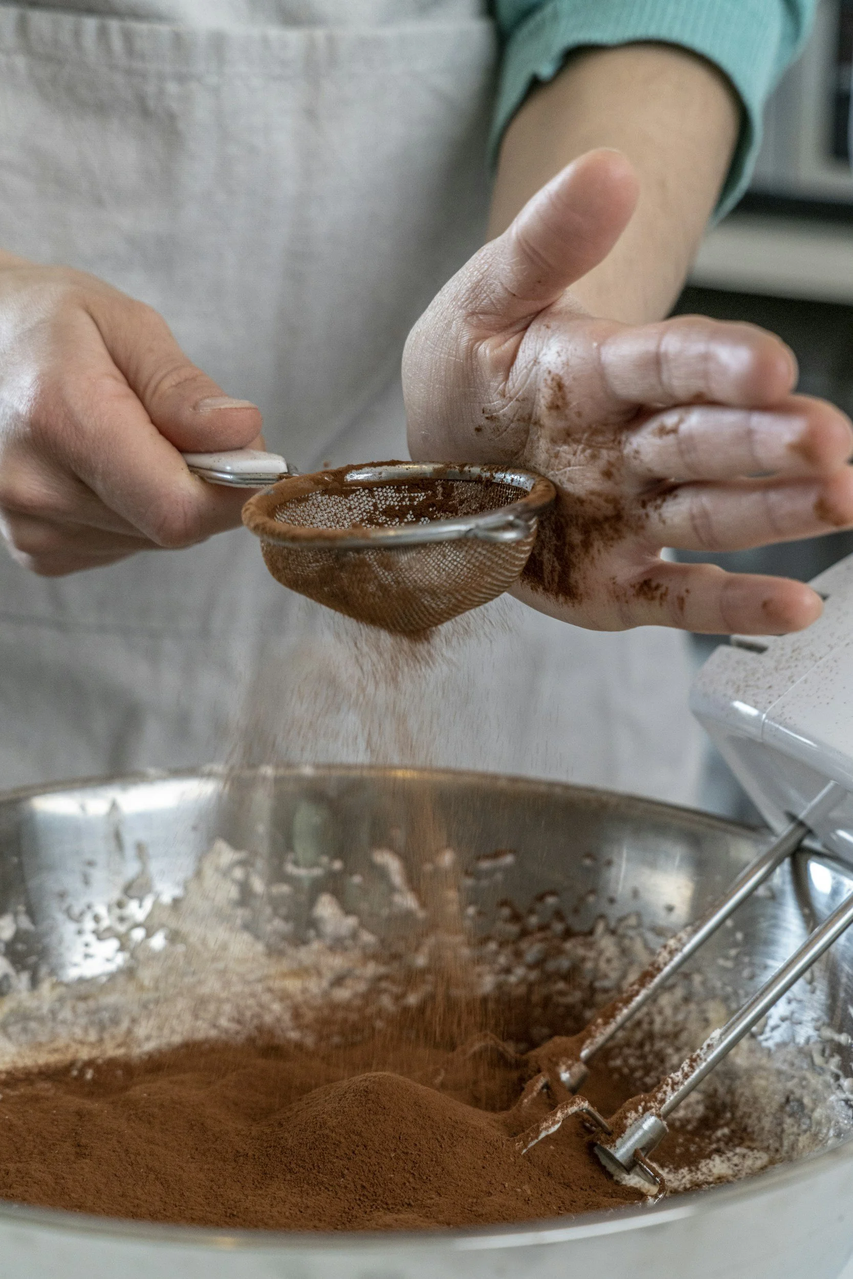 Person sifting cocoa powder into a mixing bowl with beaters, preparing ingredients for baking.