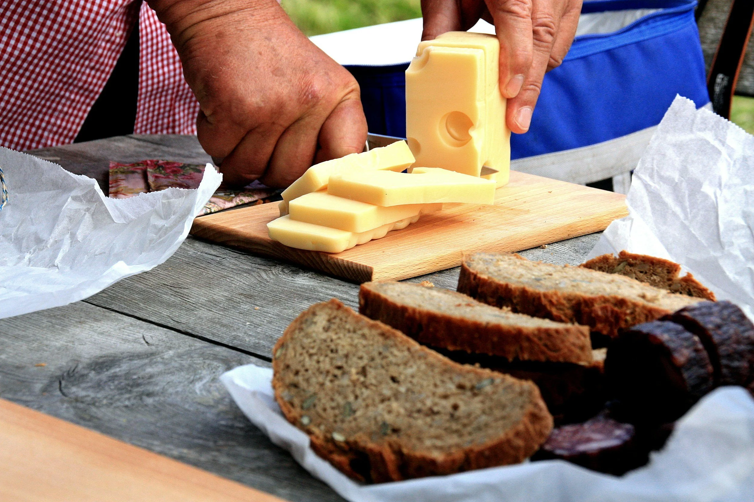 Person slicing cheese on a wooden board with rye bread and salami slices nearby.