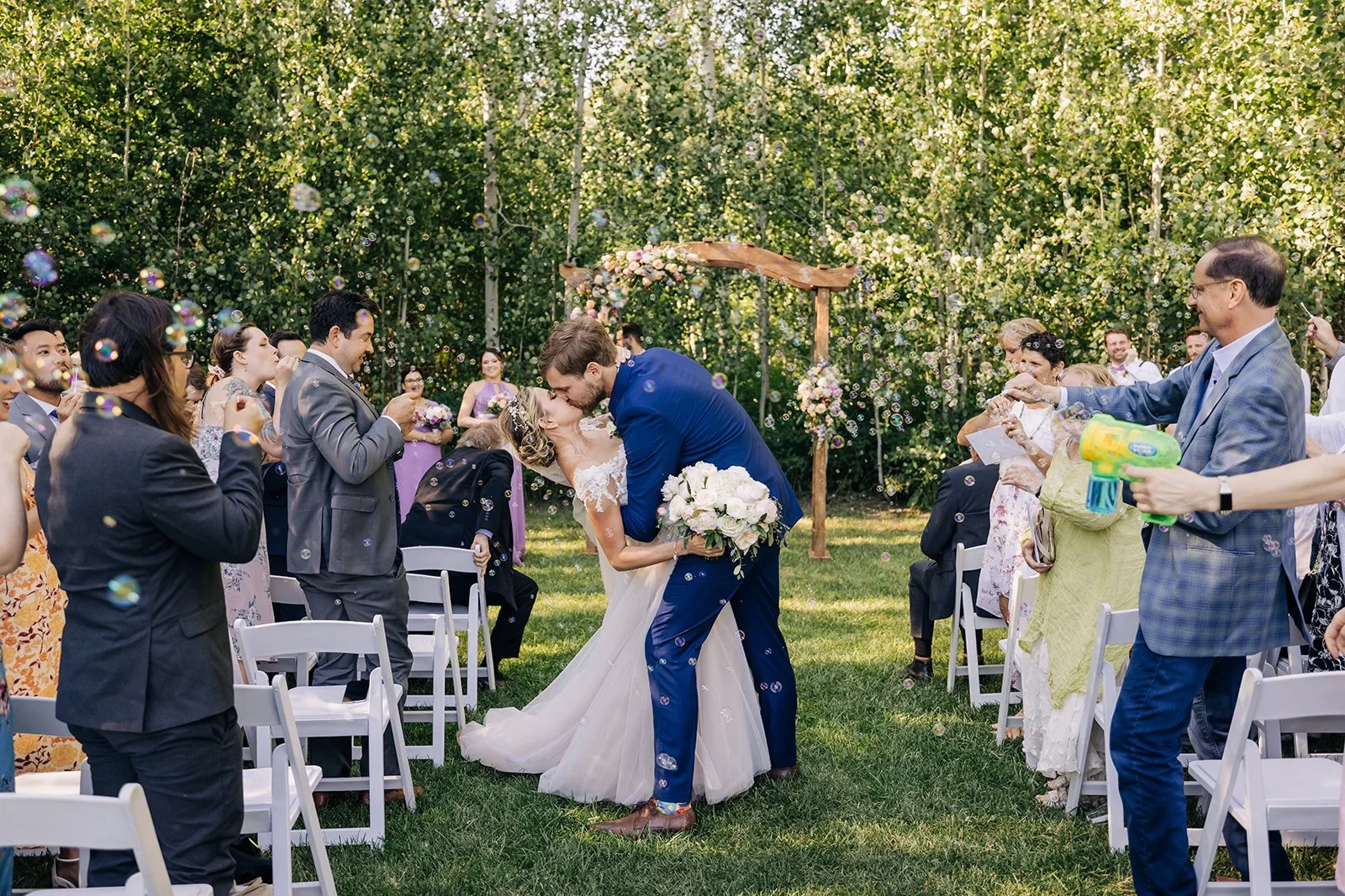 A bride and groom kissing during an outdoor wedding ceremony, surrounded by guests throwing bubbles. The scene takes place in a lush garden with green trees and floral decorations.