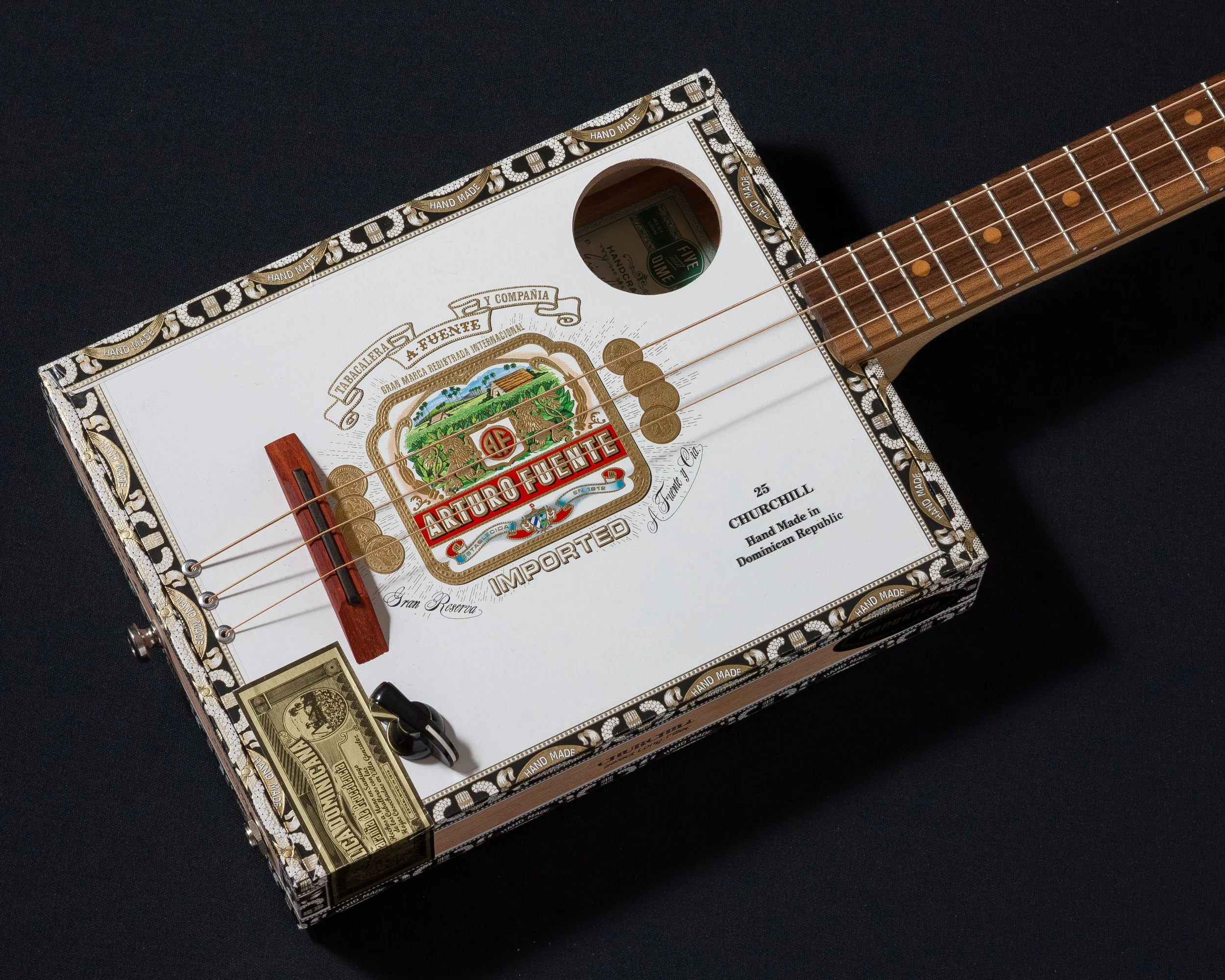 A guitar made from a Brick House cigar box, featuring four strings, a wooden bridge, and a circular sound hole on a black background. The box has decorative patterns and "Handmade in Nicaragua" inscribed on it.