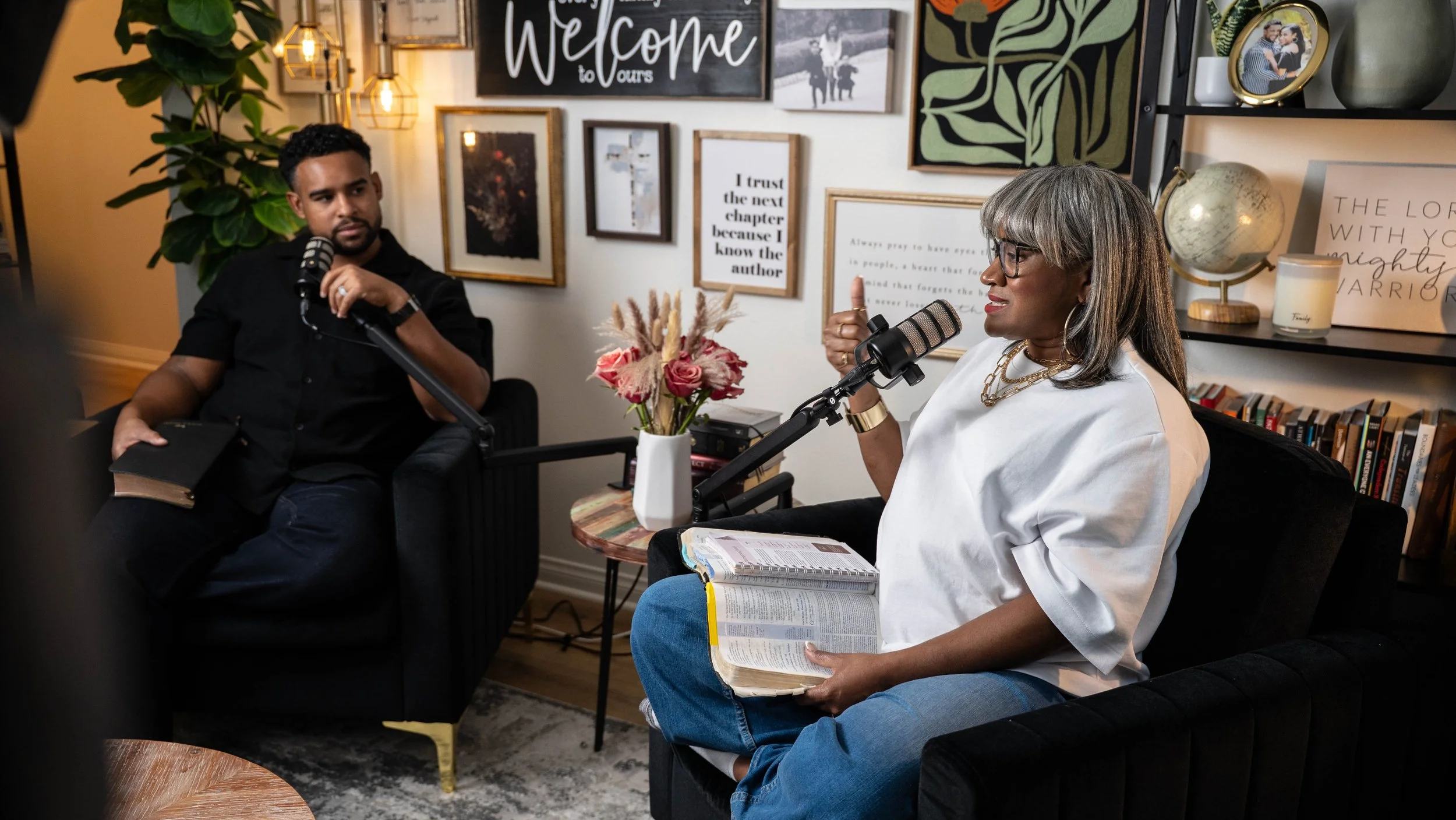 Two women engaged in a conversation in a cozy living room, with one woman speaking into a microphone and holding a Bible and notebook, while the other woman listens attentively. The room is decorated with framed pictures and quotes on the wall, a vase of pink roses, and a bookshelf with books and decorative items.