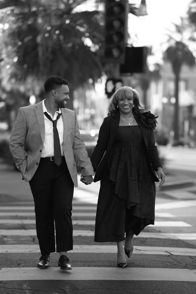 A smiling African American woman and a young African American man holding hands and crossing a city street, dressed in business attire, with palm trees and traffic lights in the background.