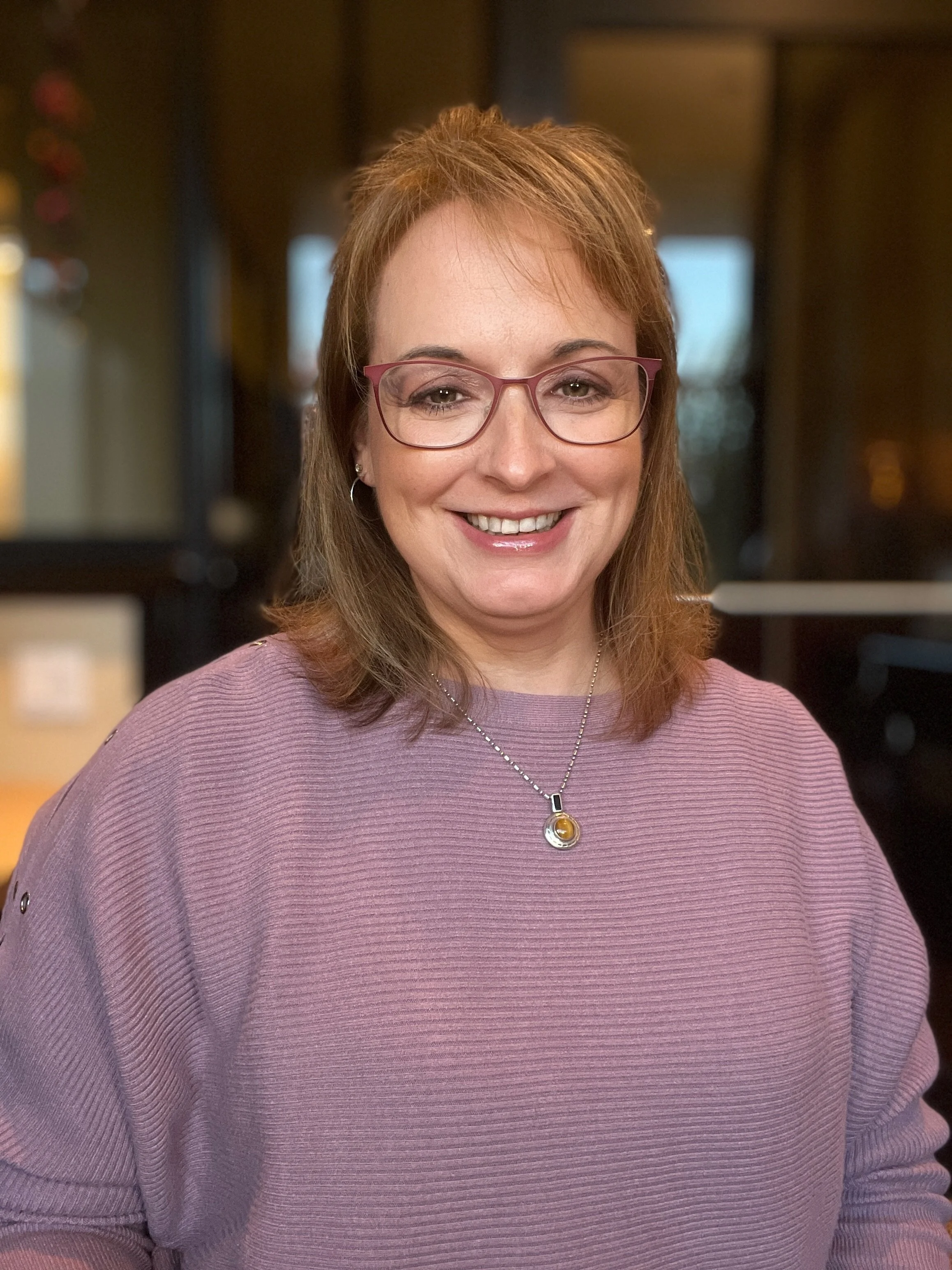 A woman with shoulder-length reddish-brown hair, pink glasses, and earrings, smiling, wearing a lavender ribbed sweater and a necklace with a circular pendant, in an indoor setting with warm lighting.