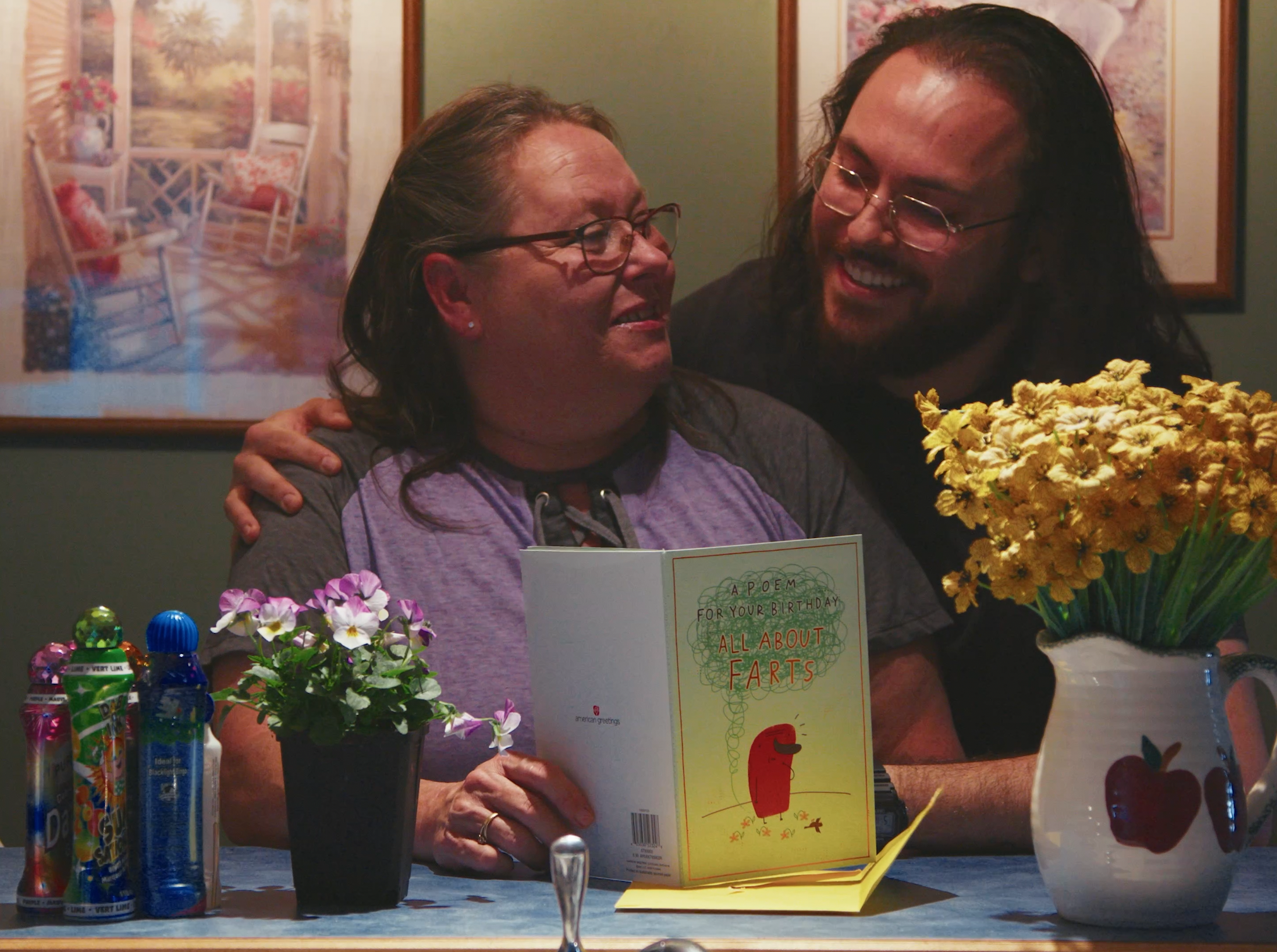 A woman holding a birthday card that reads 'APOEM FOR YOUR BIRTHDAY ALL ABOUT FARTS' while smiling. She is sitting at a table with colorful bottles and flowers, and a man with long hair and glasses is standing behind her with his arm around her shoulders. There are decorative pictures on the wall behind them.