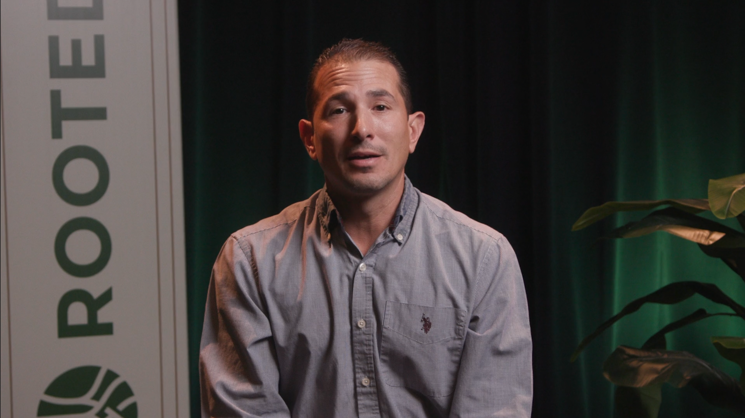 A man wearing a light gray button-up shirt, sitting indoors with a background featuring green curtains and a large plant. Part of a sign with green text is visible on the left side.
