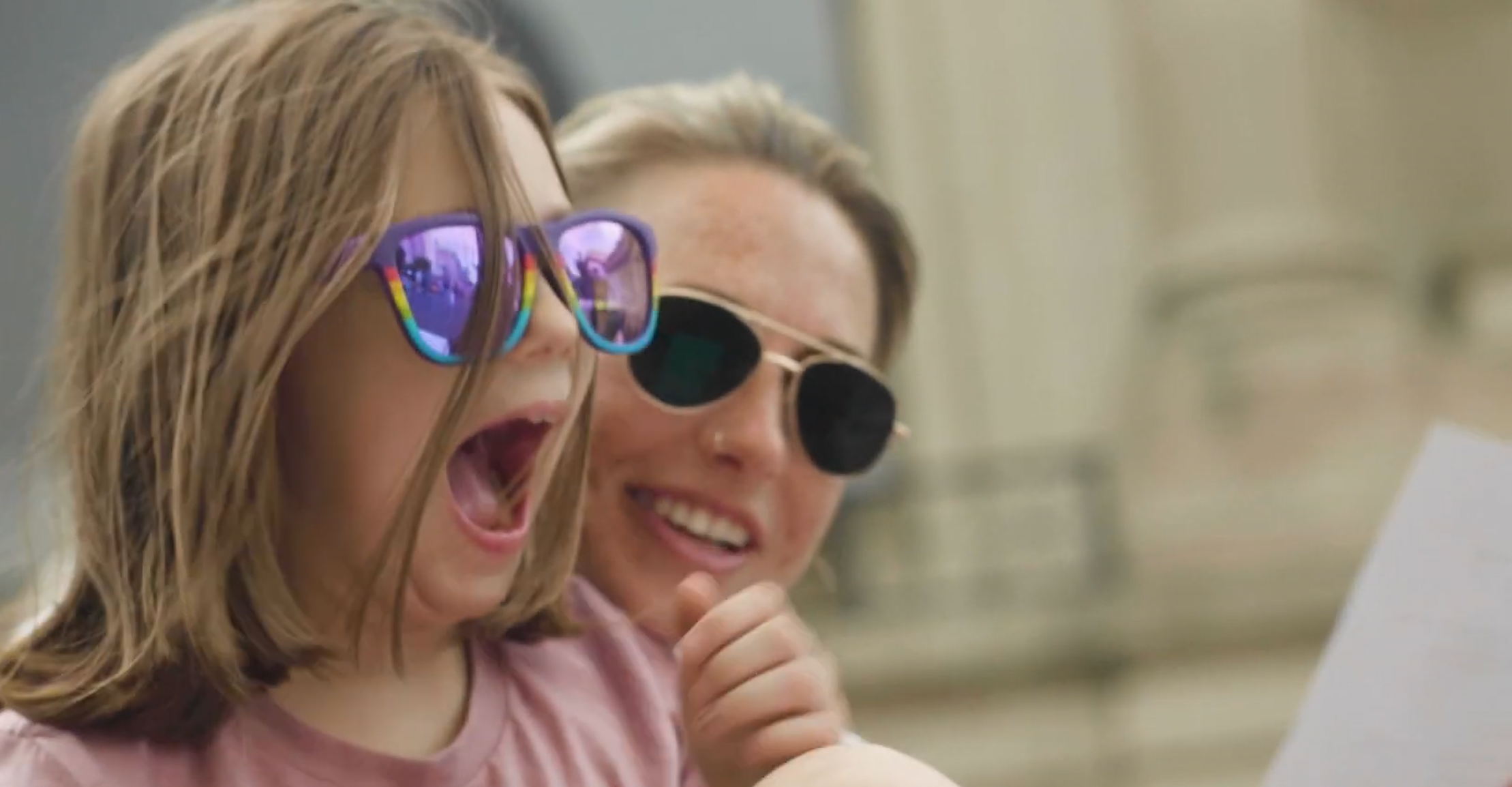 Two women, both wearing sunglasses, look at a tablet together. The woman on the left has long wavy hair and is wearing reflective sunglasses, while the woman on the right has her hair in an updo and wears round black sunglasses. They appear happy and engaged, possibly looking at something amusing on the device.