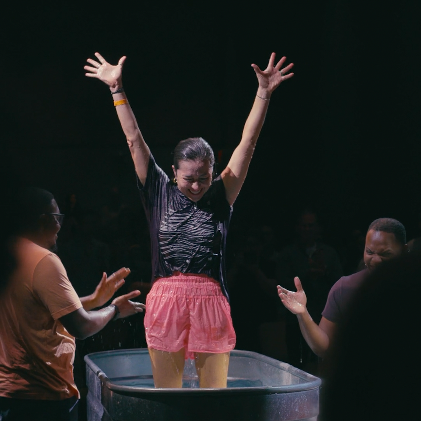 A woman in a patterned black shirt and pink shorts with her arms raised and smiling, standing in a water basin surrounded by people clapping and cheering.