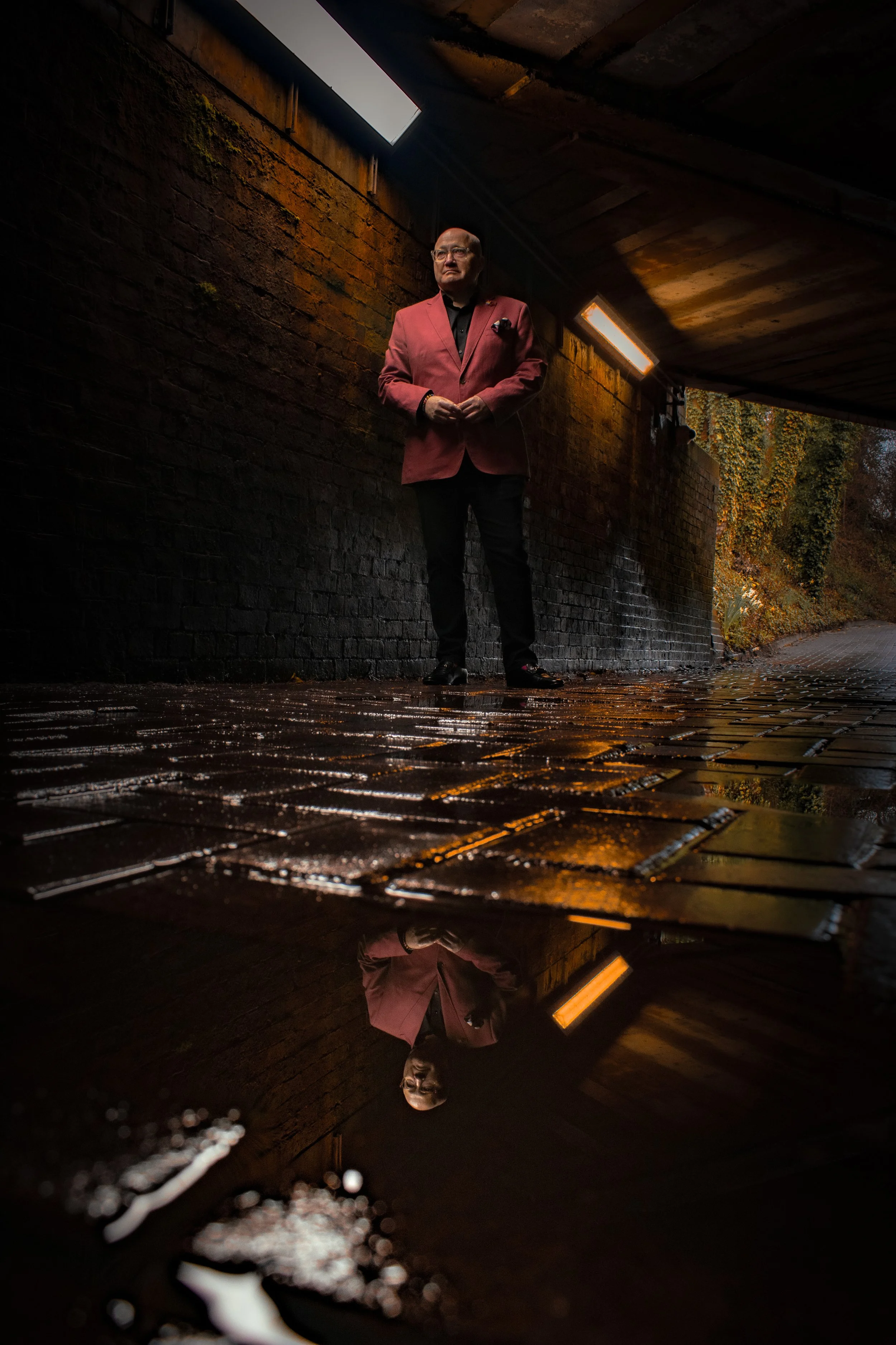 A man in a pink blazer and black pants standing under an overpass, with wet brick pavement reflecting his image.