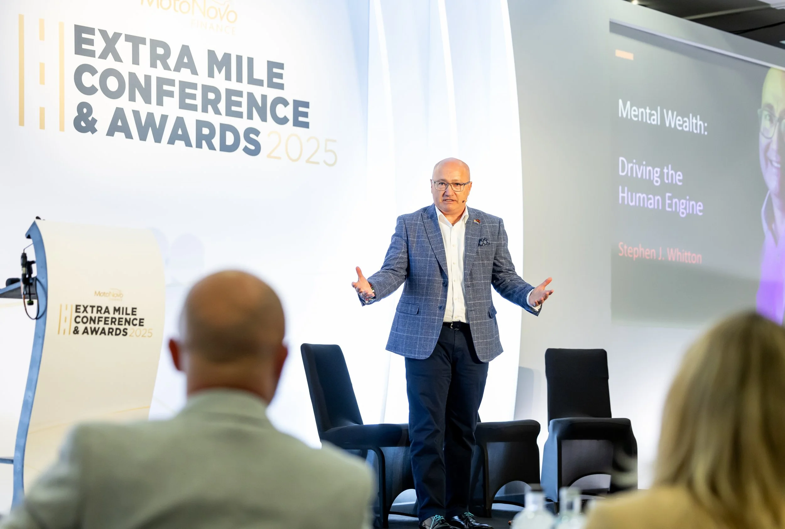 A man in a gray checked blazer and glasses giving a presentation at the Extra Mile Conference & Awards 2025, with a large screen displaying the title 'Mental Wealth: Driving the Human Engine' and the speaker's name 'Stephen J. Whitton'.