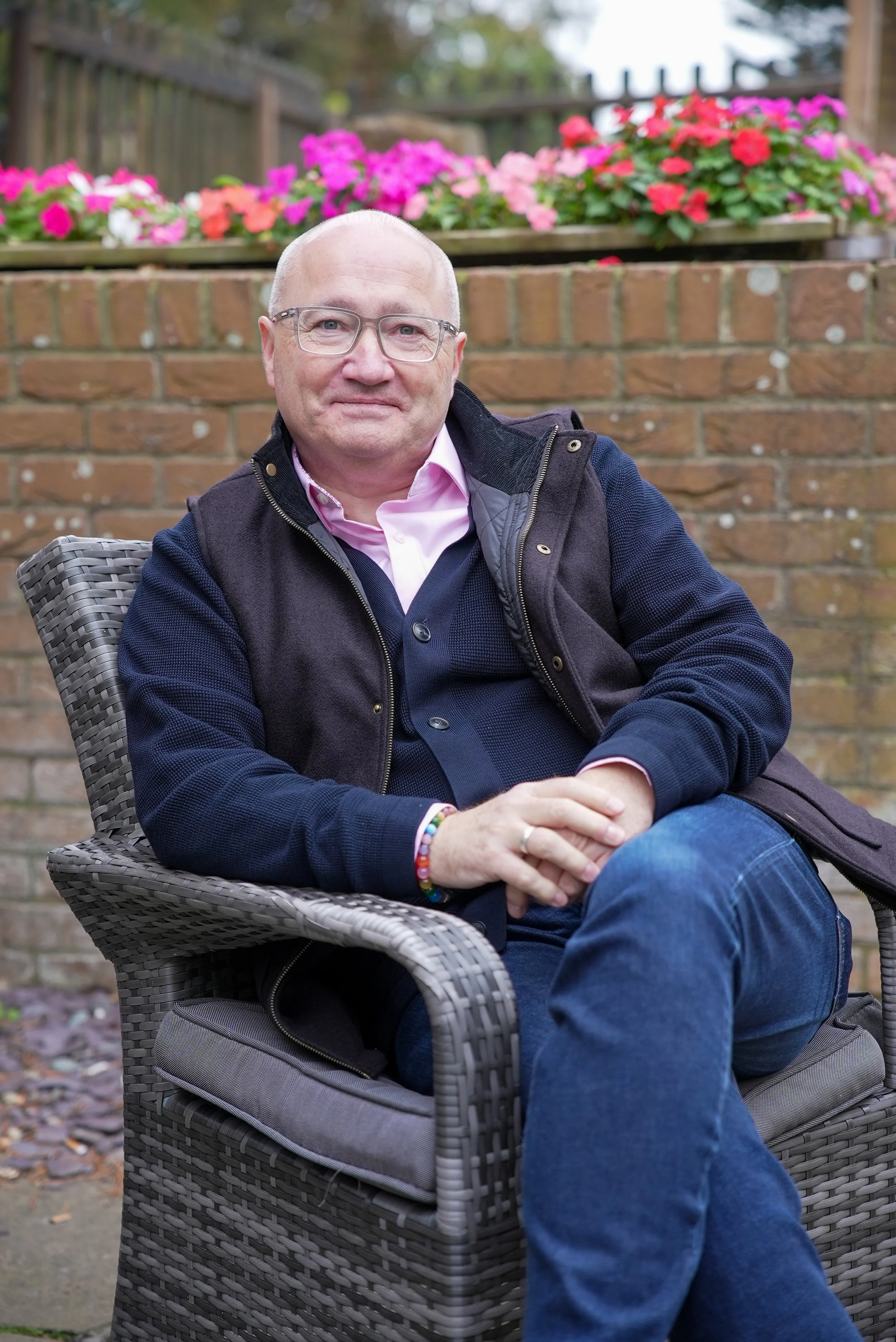 A middle-aged man with glasses and a light pink shirt sitting outdoors on a wicker chair, smiling with hands folded on his lap, in front of a brick wall and colorful flowers in the background.