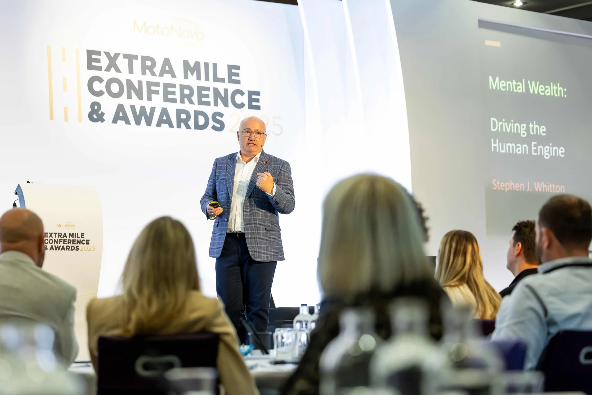 A man giving a presentation at the Extra Mile Conference & Awards 2025. The screen displays the title 'Mental Wealth: Driving the Human Engine' by Stephen J. Whitton. Audience members are seated and listening.