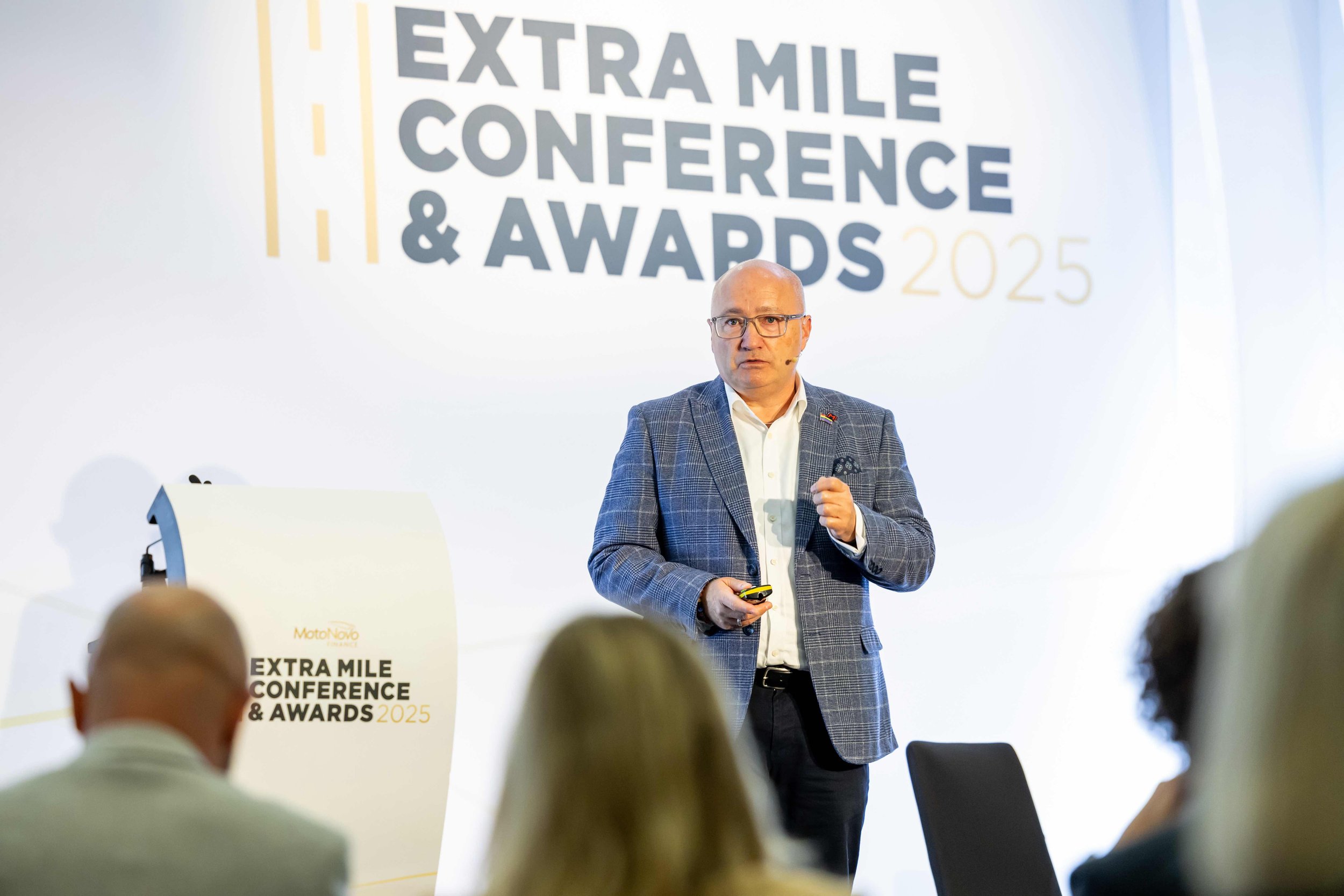 A man in a plaid suit jacket and glasses giving a presentation at the Extra Mile Conference & Awards 2025, standing in front of a white backdrop with the event name.