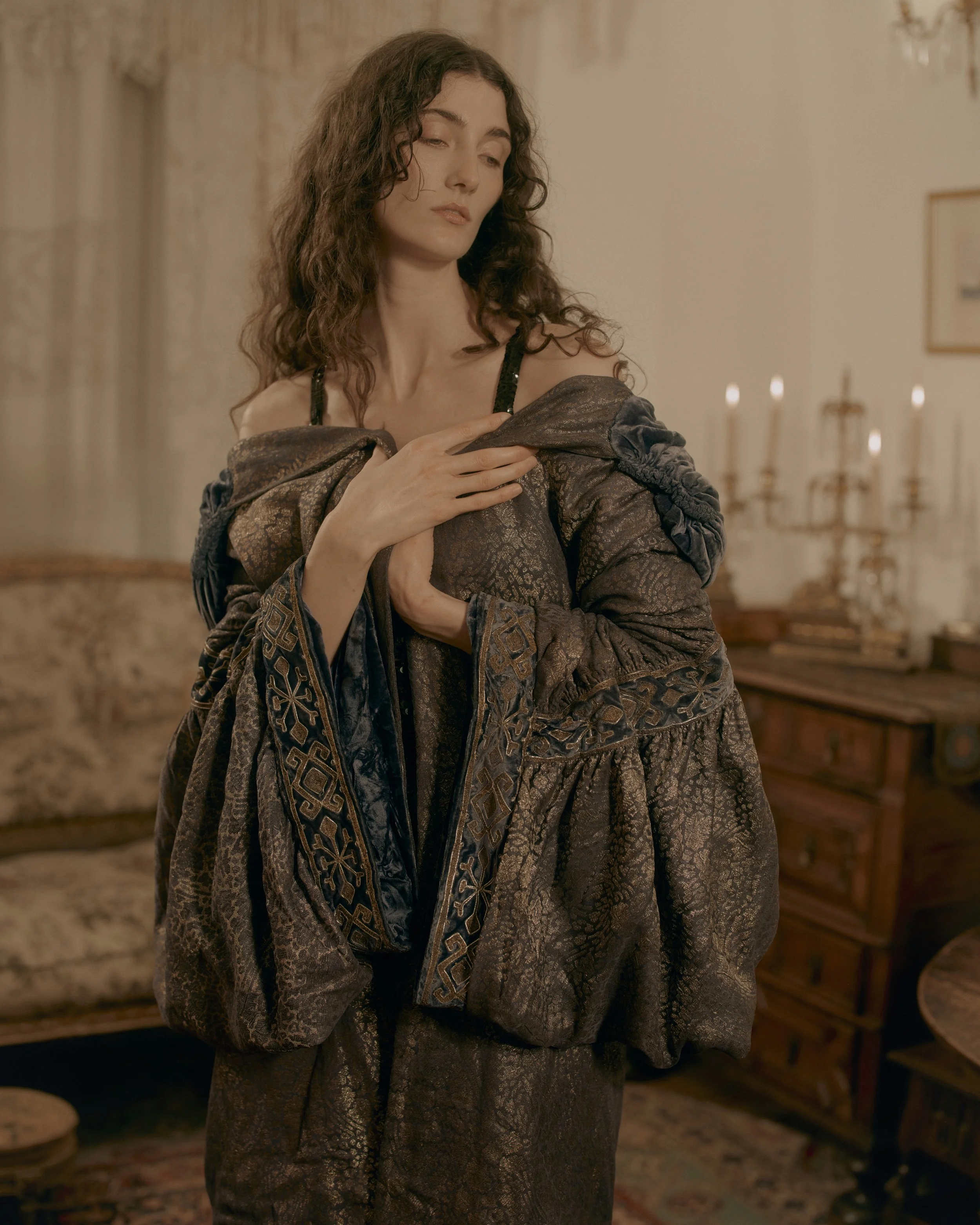 A woman in a vintage, ornate brown gown with intricate embroidery, standing with her eyes closed and her hand on her chest, in a room with candlelit background and antique furnishings.