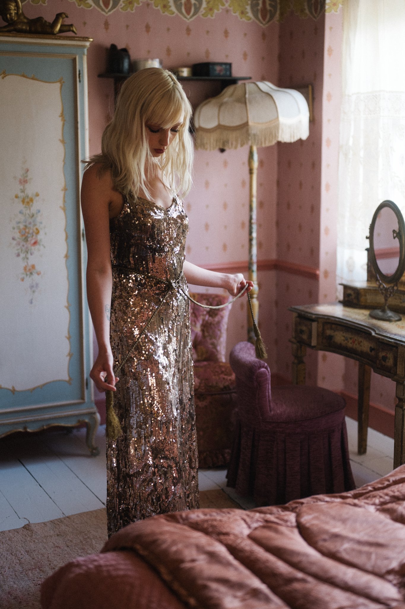 A woman with blonde hair in a glittering gold dress standing in vintage-style bedroom, holding a decorative tassel.