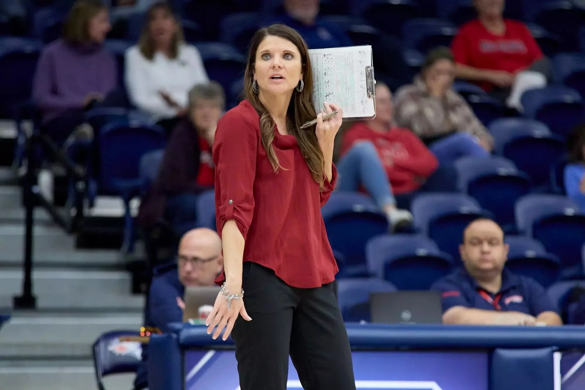 Close-up of a smiling woman with curly hair, glasses, hoop earrings, and layered necklaces, in an indoor setting.