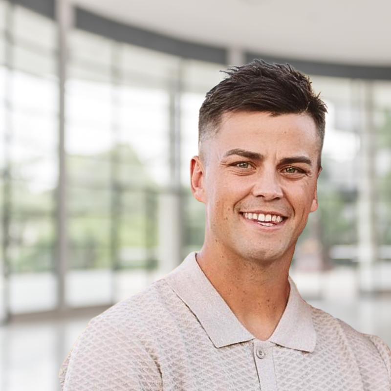Smiling young man with short dark hair, wearing a light-colored collared shirt, standing inside a modern building with large glass windows.