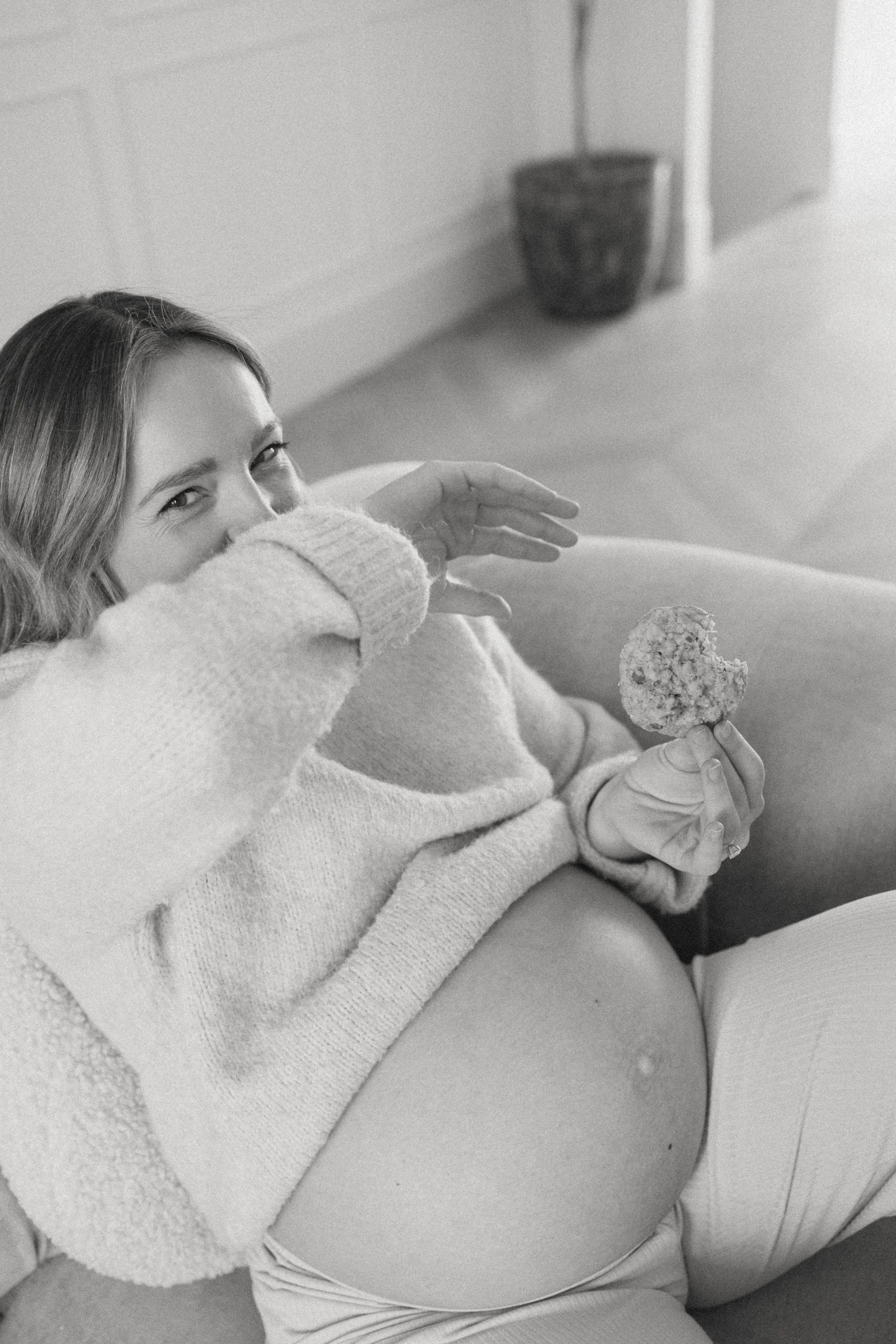 Pregnant woman sitting on a sofa, covering her face with one hand while holding a cookie in the other.