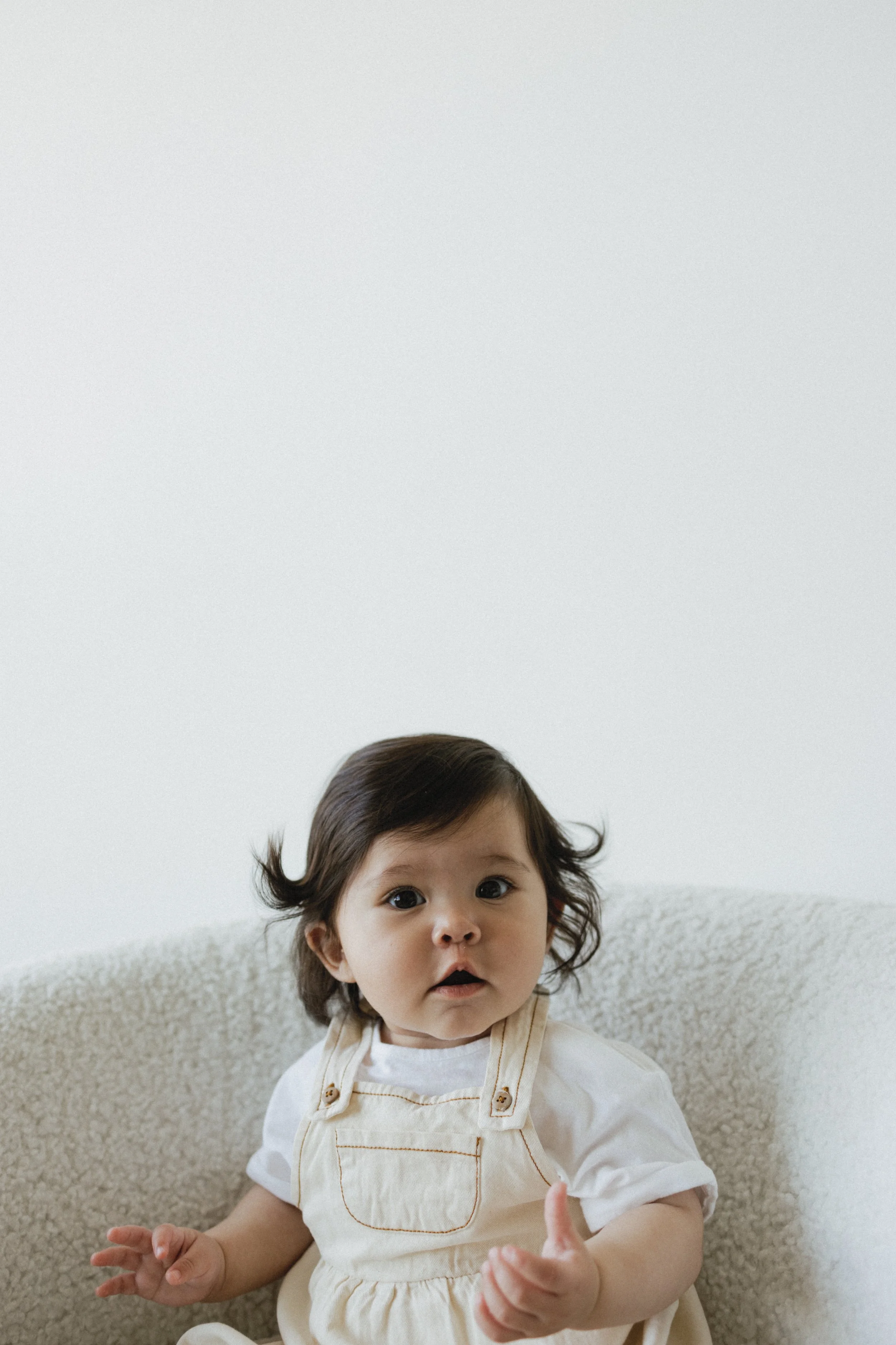 Baby sitting in a fluffy chair wearing a white shirt and beige overalls, looking forward.
