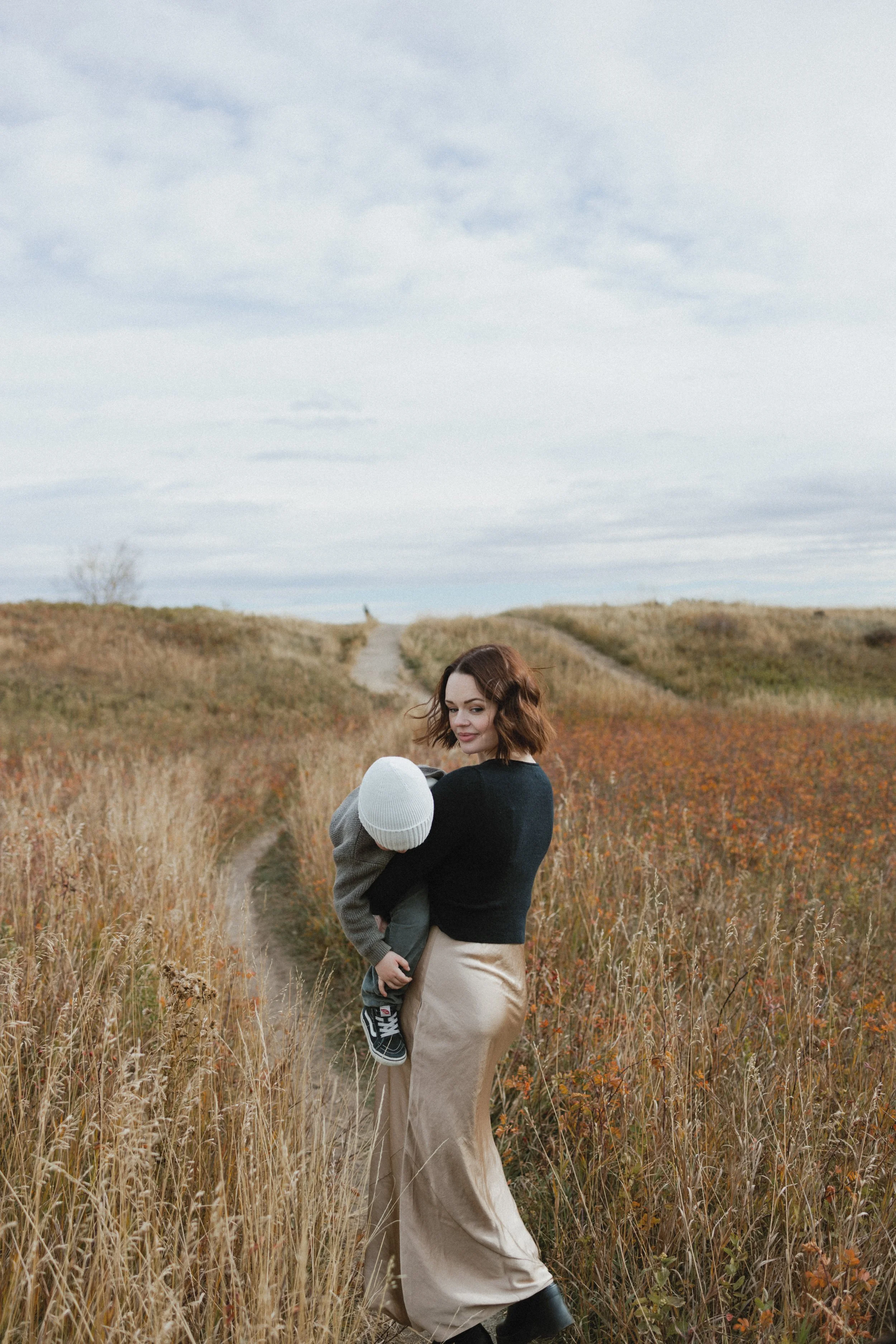 Woman carrying a child in a field with tall grass under a cloudy sky.