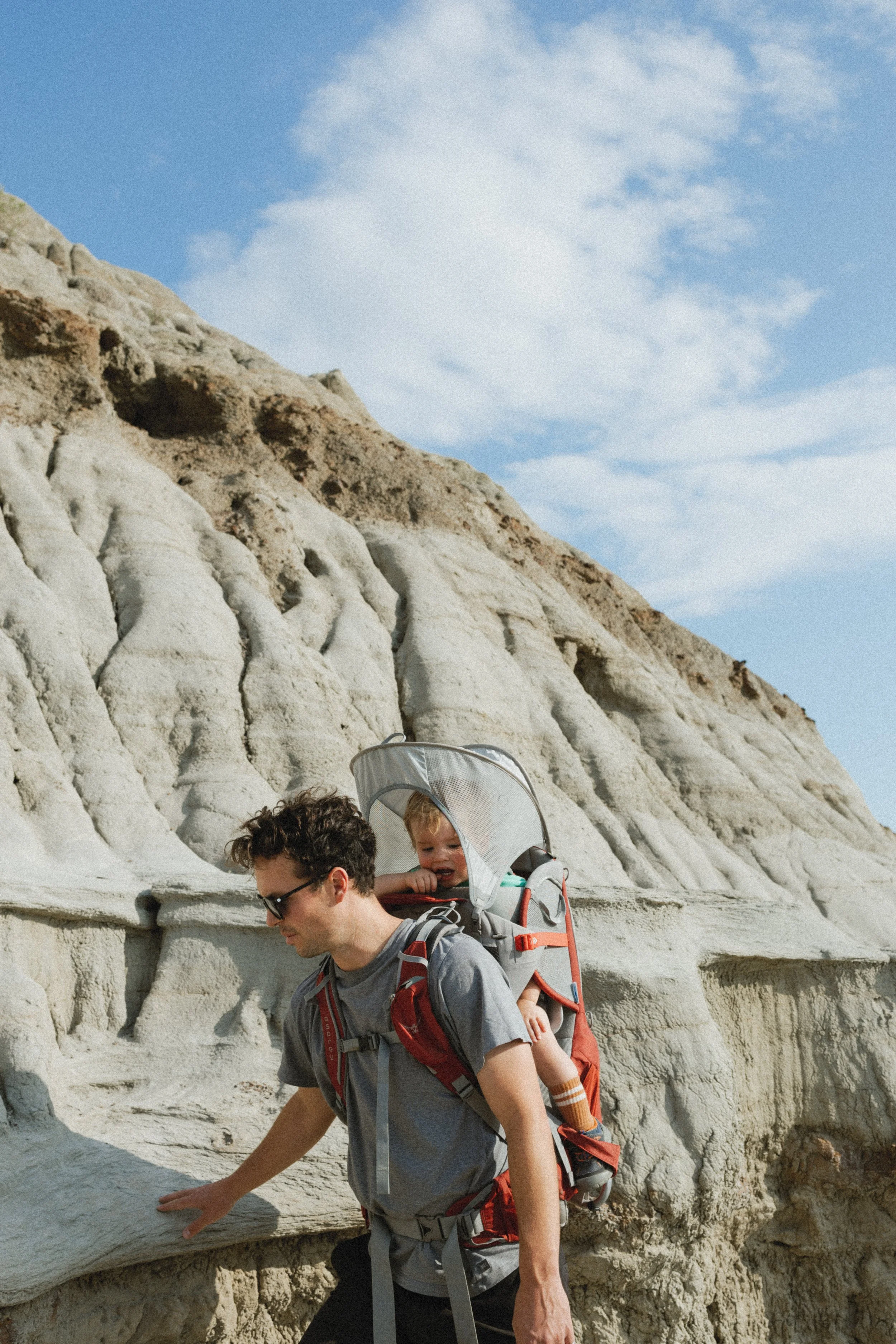Man hiking with a baby carrier on a rocky landscape under a clear sky.