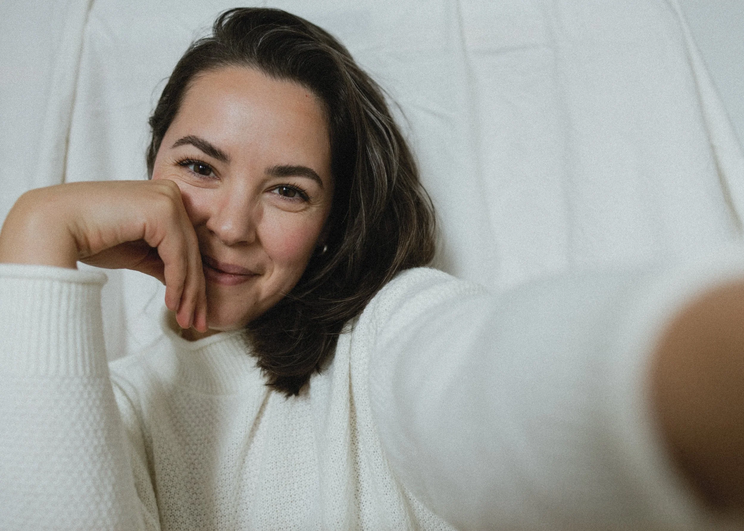 Person smiling, wearing a white sweater, taking a selfie against a plain background.