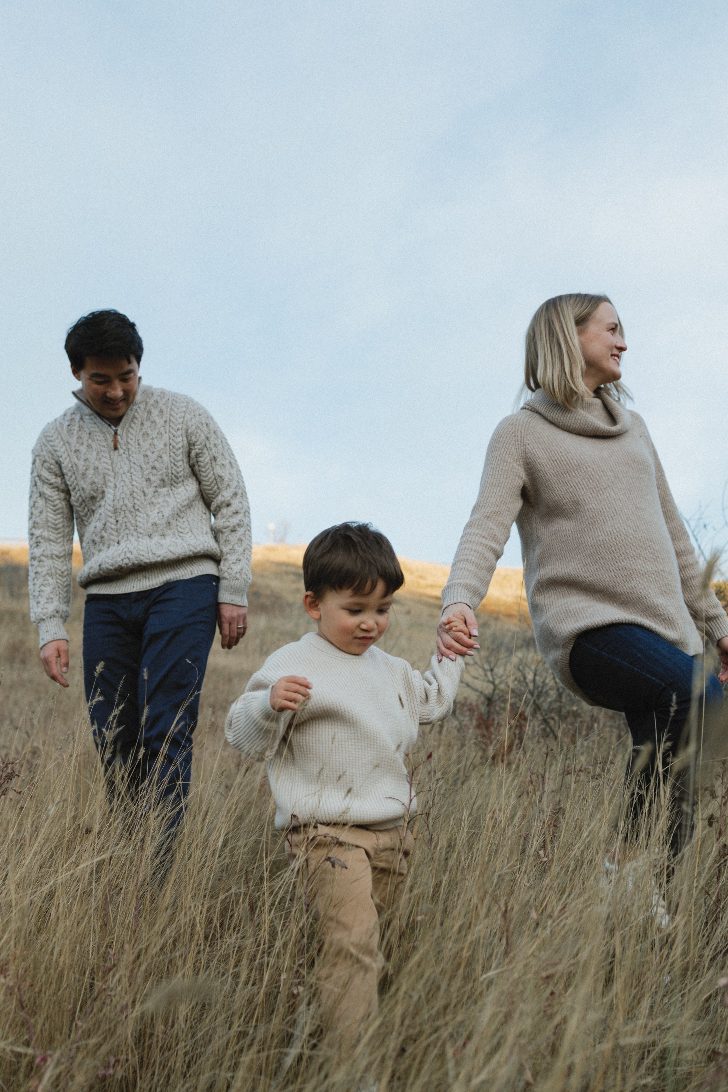 Family walking through a field wearing sweaters, with a child holding an adult's hand.