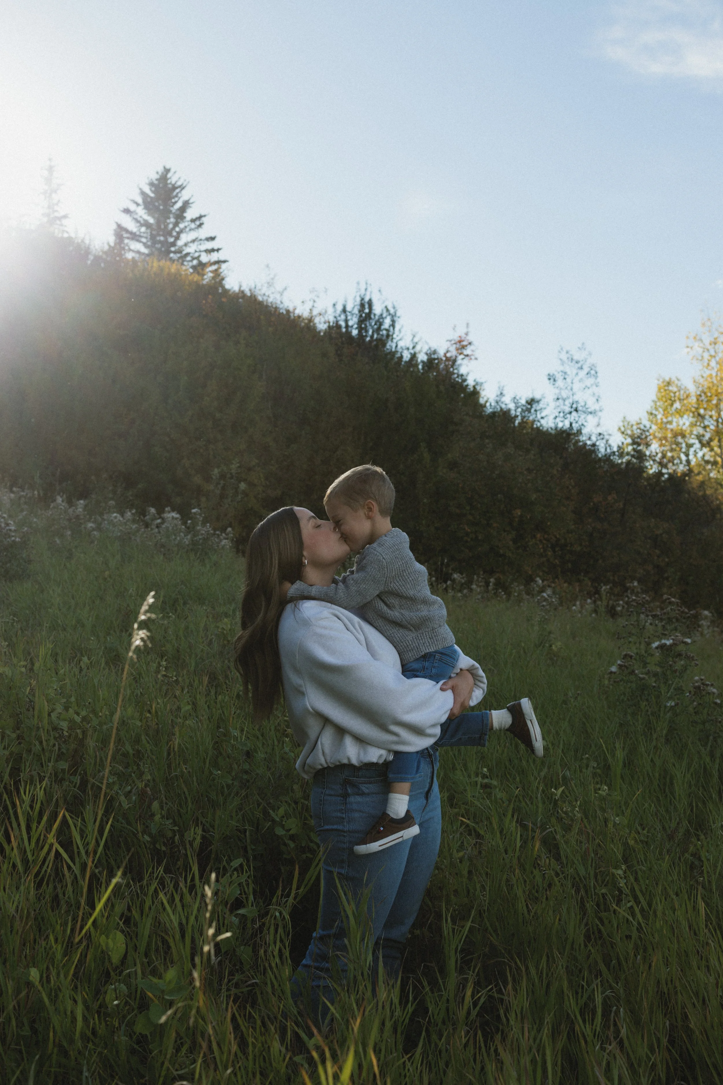 A young woman embraces a child in a grassy field with trees in the background, as sunlight filters through the trees.