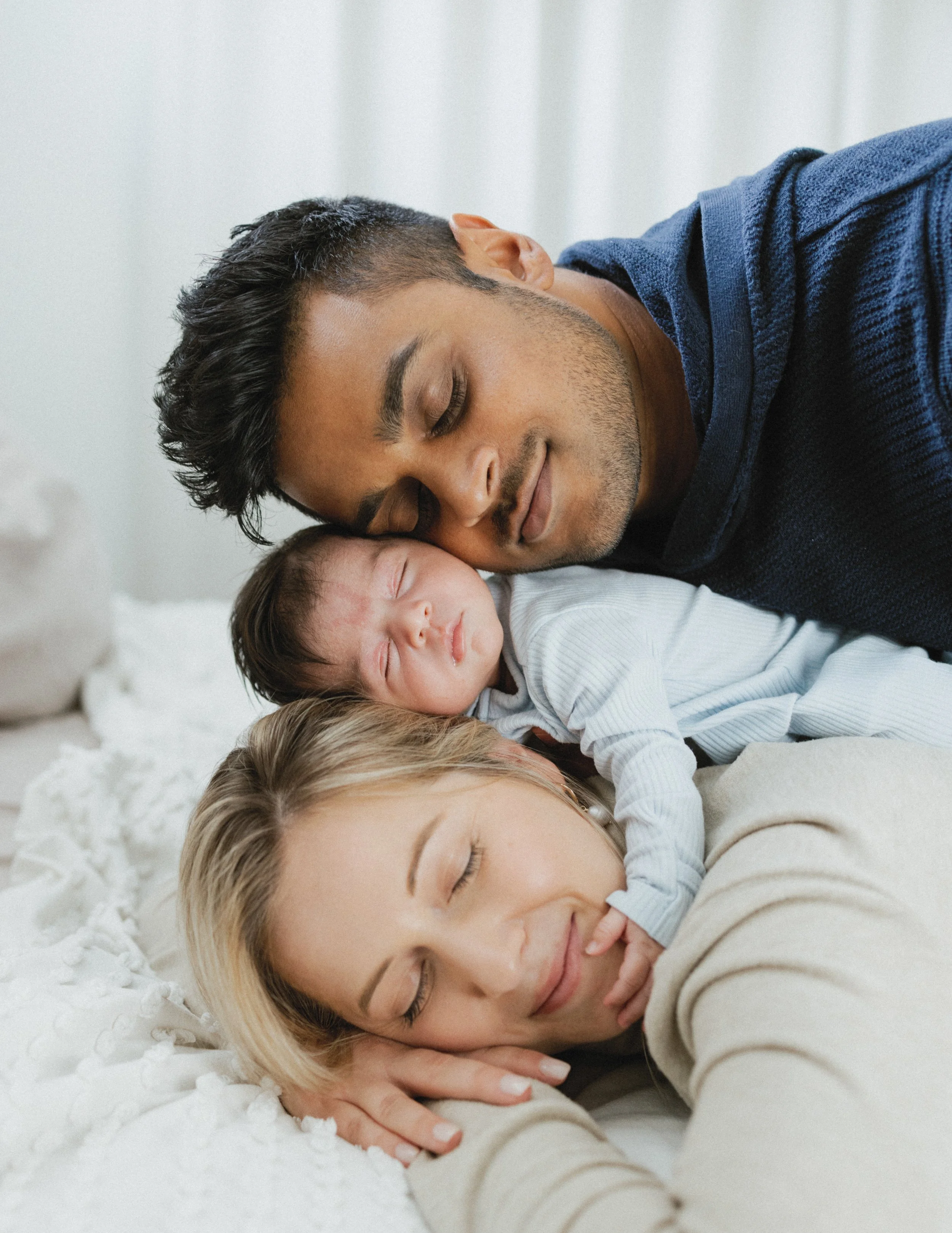 Family with baby lying together on a bed, smiling and relaxed.