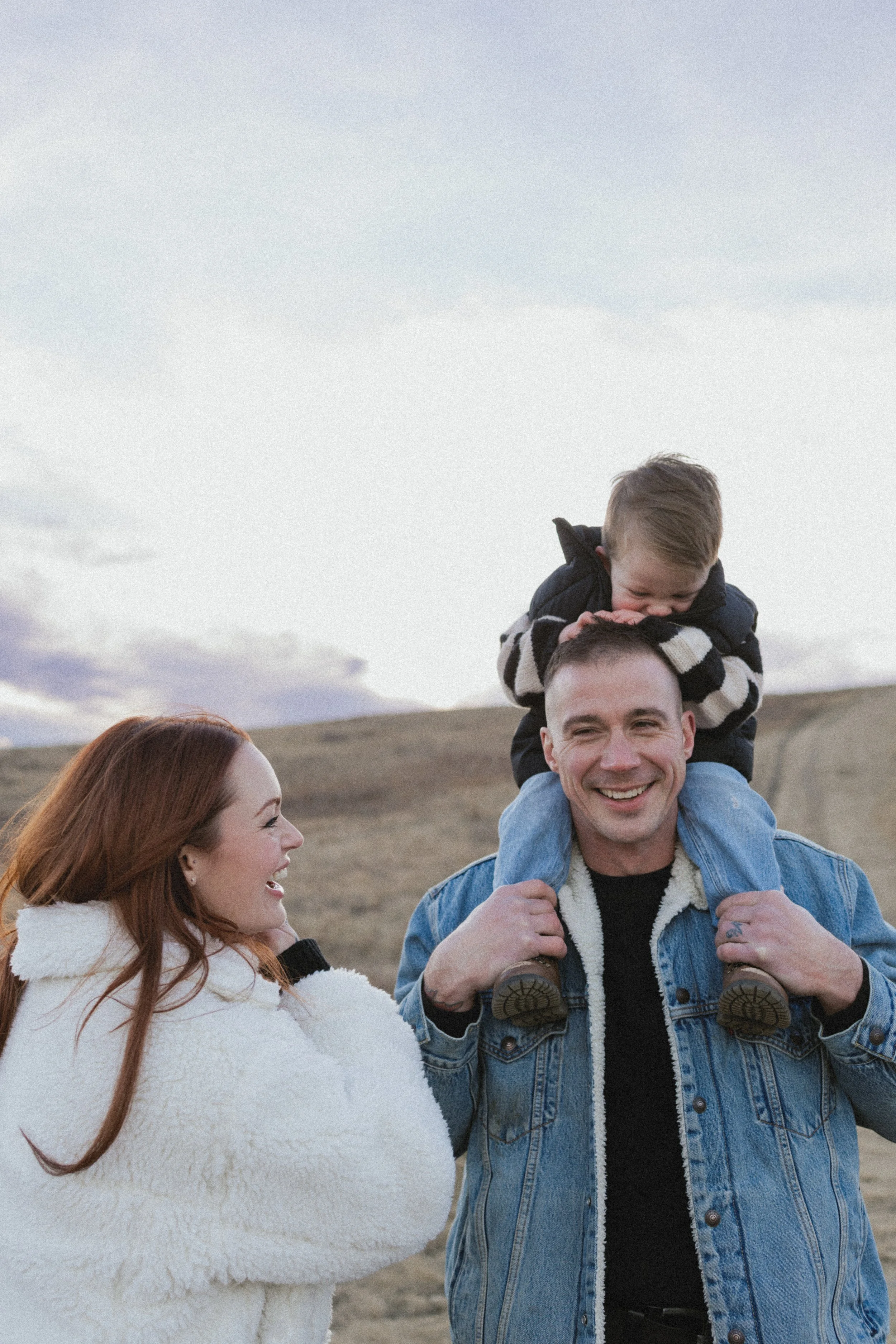 A family enjoying an outdoor moment, with a child sitting on a man's shoulders, both wearing warm jackets. A woman in a cozy sweater looks at them, smiling. The background shows a cloudy sky and a rural landscape.