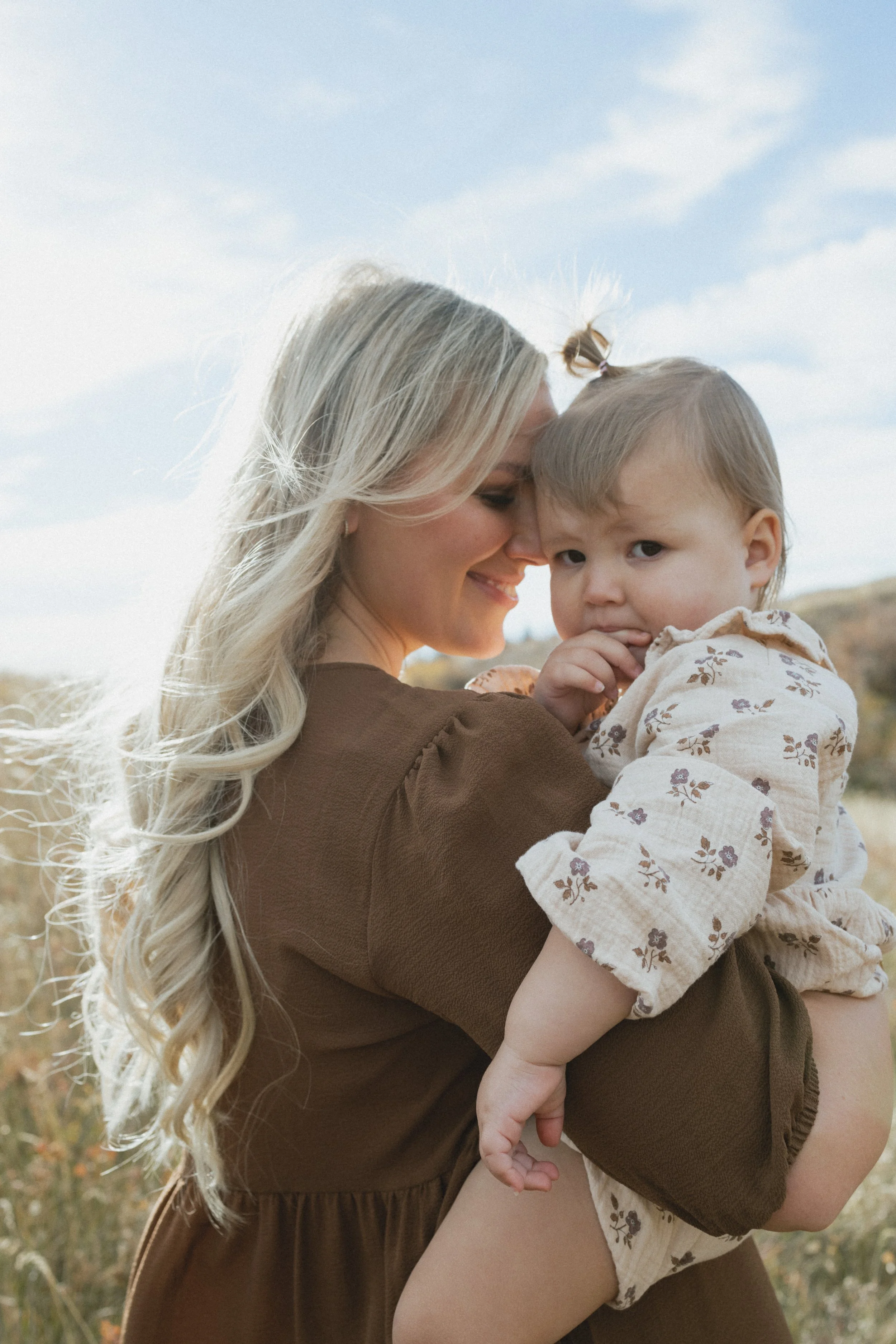 Woman holding a baby in a field, smiling, with long blonde hair and wearing a brown dress, baby dressed in floral pattern clothing, under blue sky.