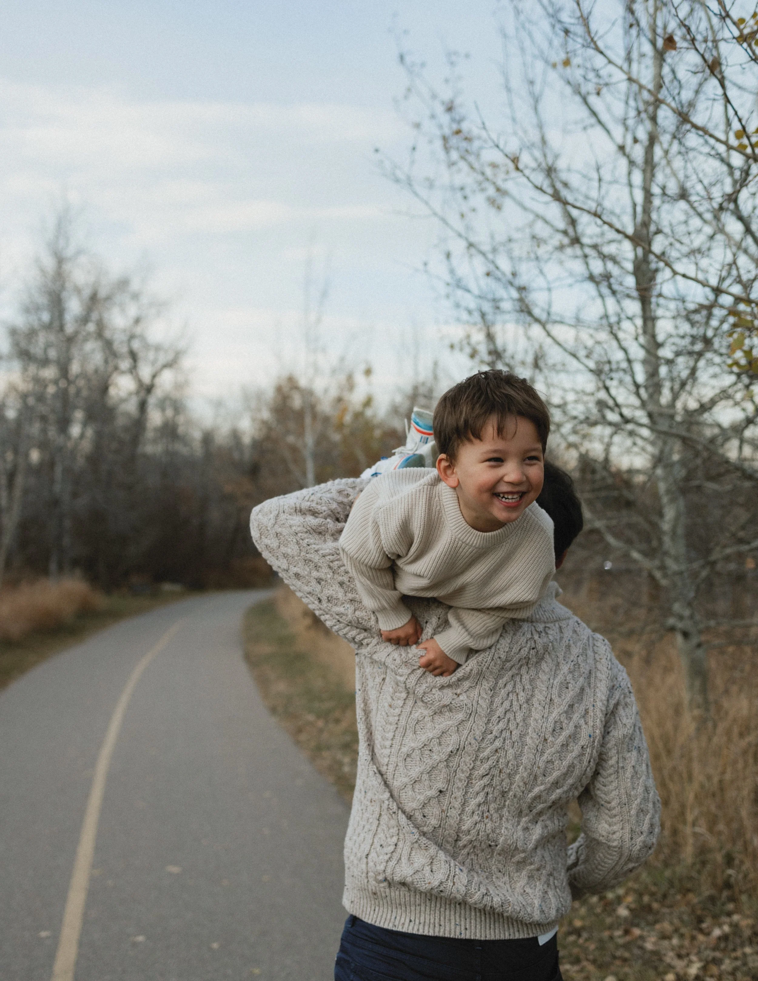 Man in a knit sweater walking on a path carrying a laughing child draped over his shoulder.